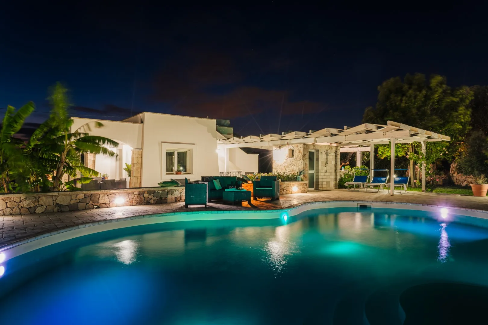 Una piscina iluminada por la noche junto a una zona de estar en el patio.
