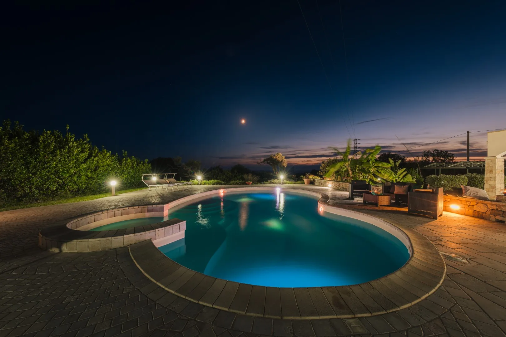 Una piscina exterior iluminada al anochecer, rodeada de pavimento y plantas de jardín.