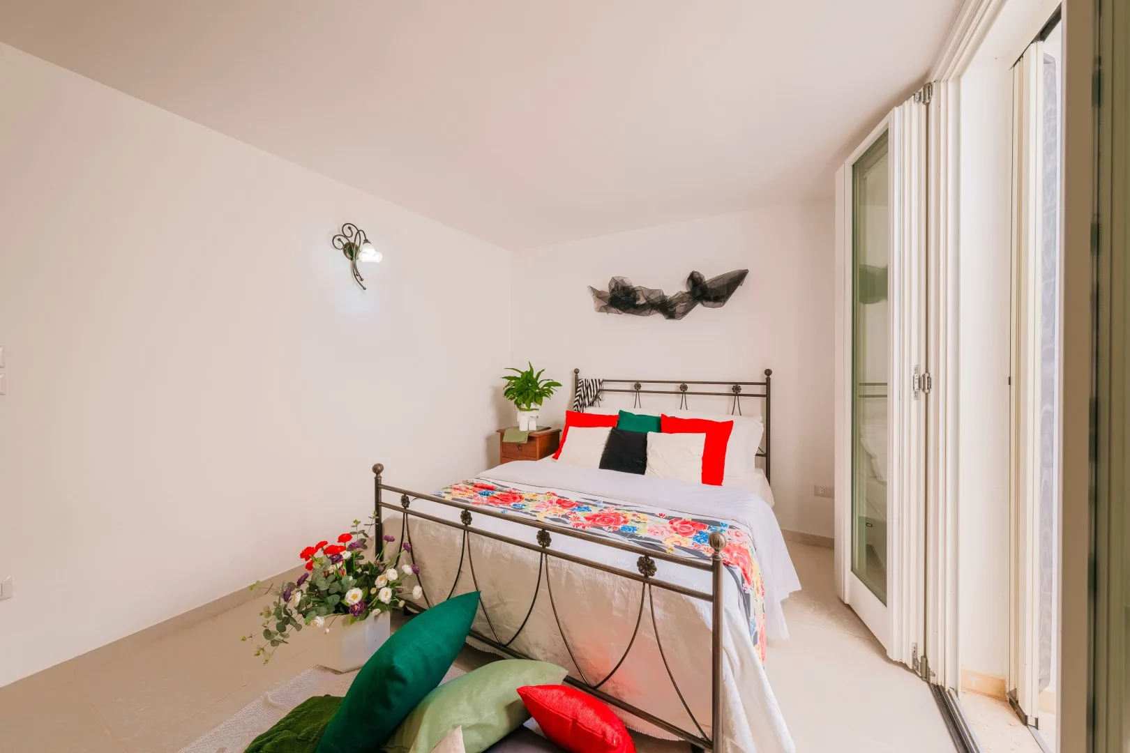 Un dormitorio luminoso con una cama de metal, ropa de cama colorida y flores al lado.