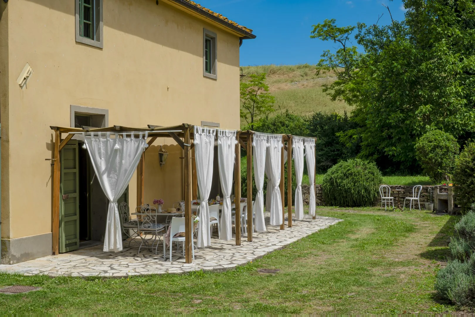 Un patio ajardinado junto a una casa tiene asientos bajo una pérgola con cortinas y un césped.
