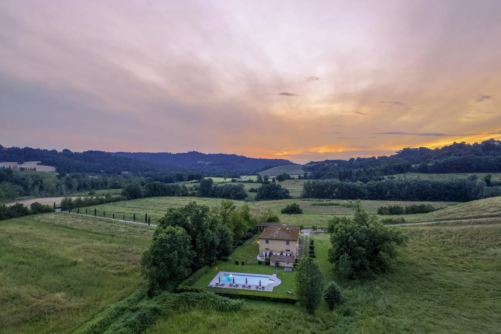 Un paisaje rural al atardecer, con una casa y una piscina entre campos verdes.