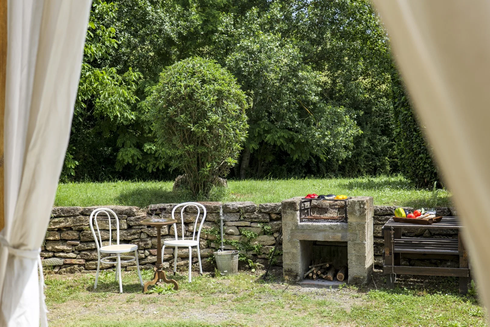 Un patio exterior con asientos y una barbacoa de piedra junto a un jardín frondoso.