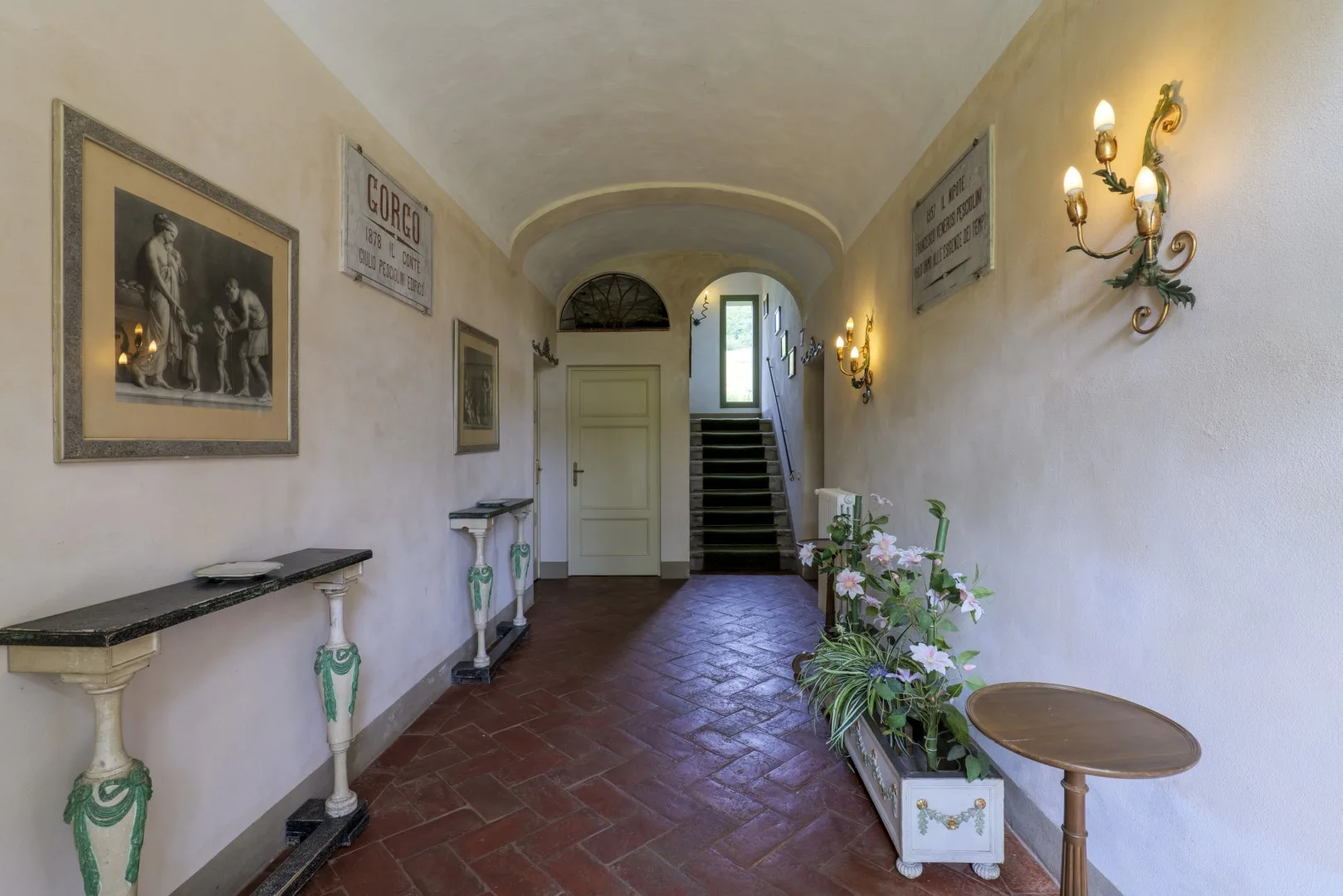 Un pasillo interior largo con cuadros, apliques de pared y plantas en macetas.