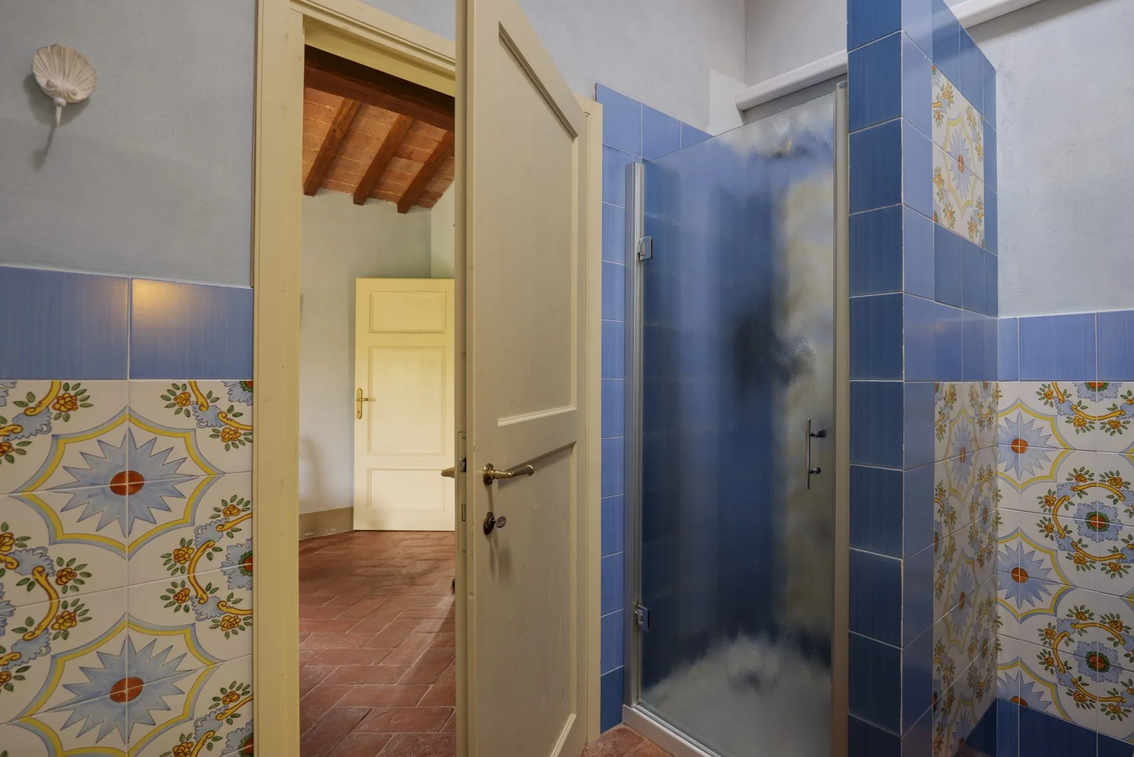 Un baño alicatado con una ducha de cristal y una puerta abierta que da a otra habitación.