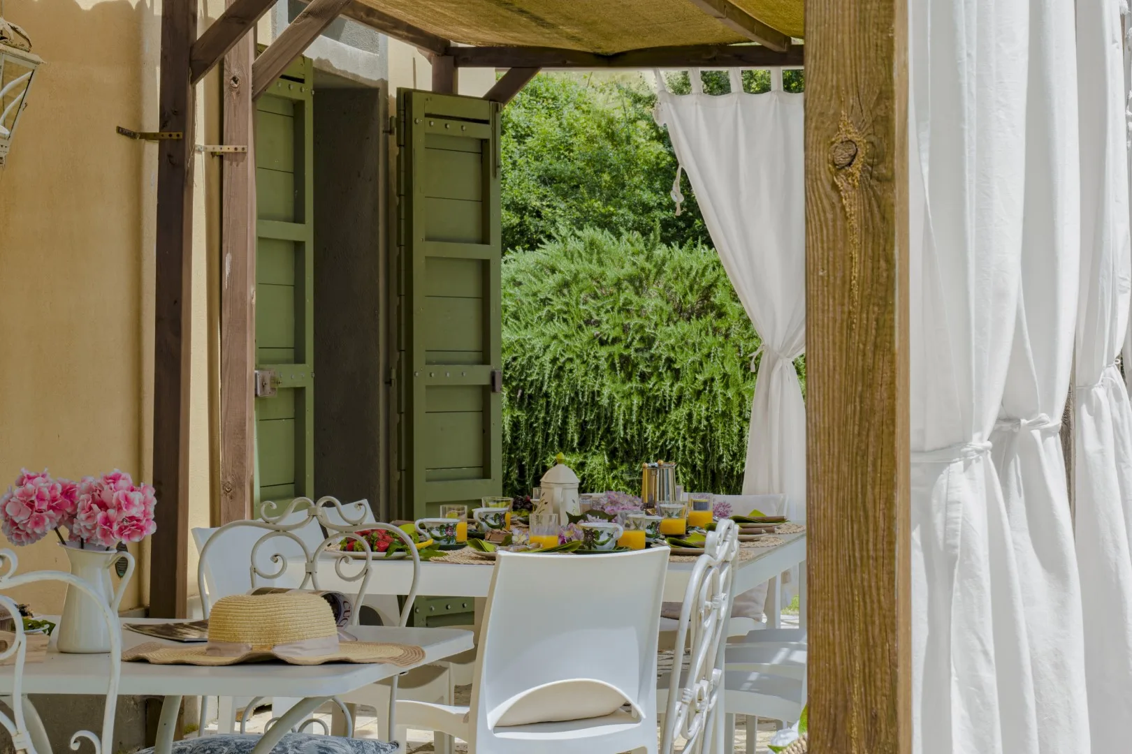 Comedor al aire libre con una mesa puesta junto a contraventanas verdes y cortinas blancas.