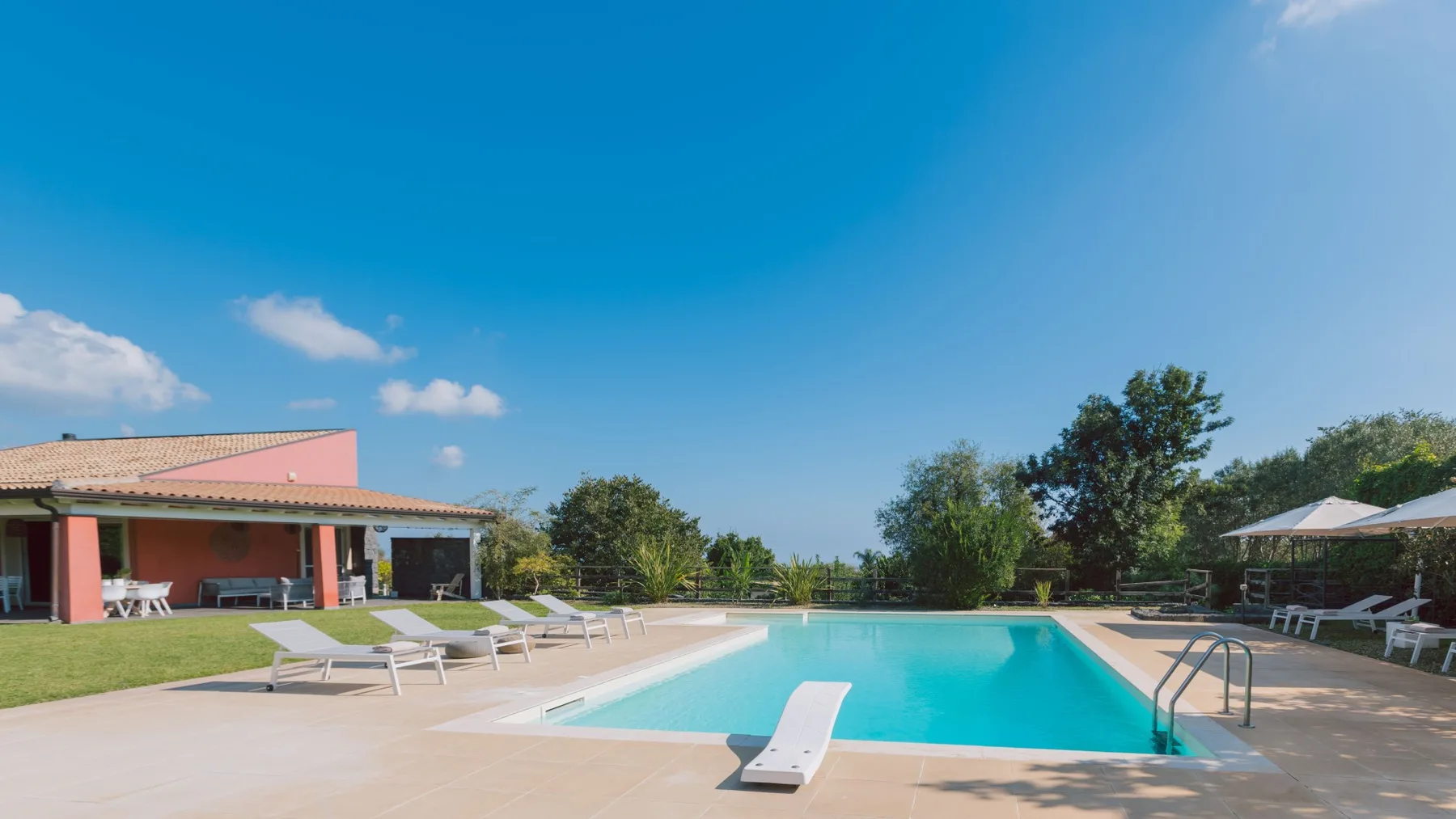 Piscina al aire libre junto a un patio, con tumbonas y vegetación.