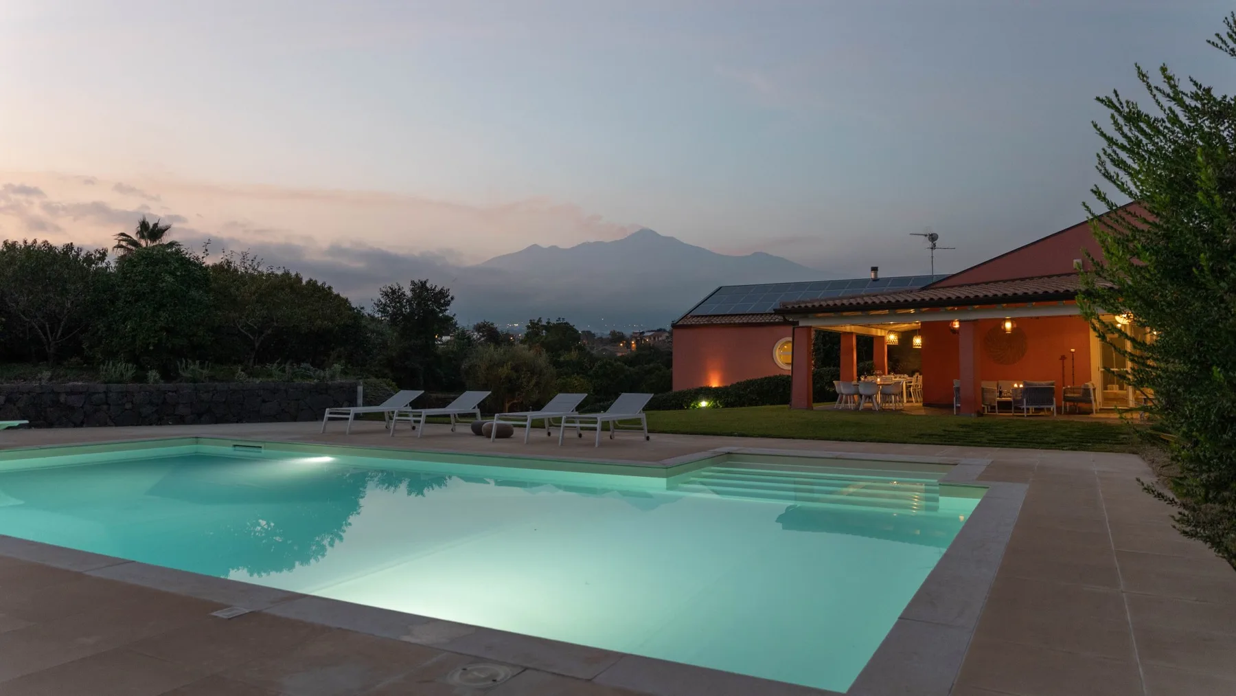Una piscina al aire libre junto a un edificio iluminado, con montañas a lo lejos.