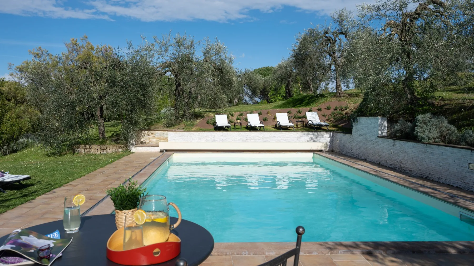 Una piscina está rodeada por un jardín con árboles y tumbonas.