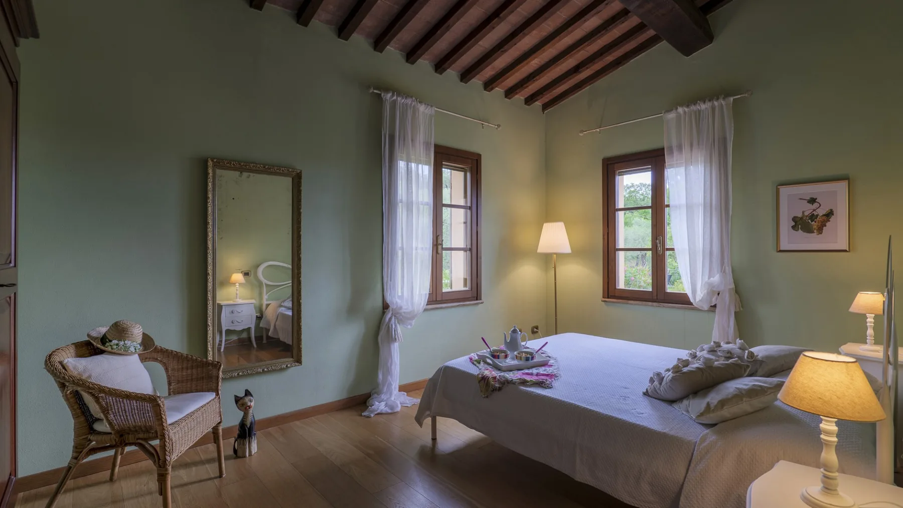Un dormitorio con una cama arreglada y ventanas altas con cortinas traslúcidas.
