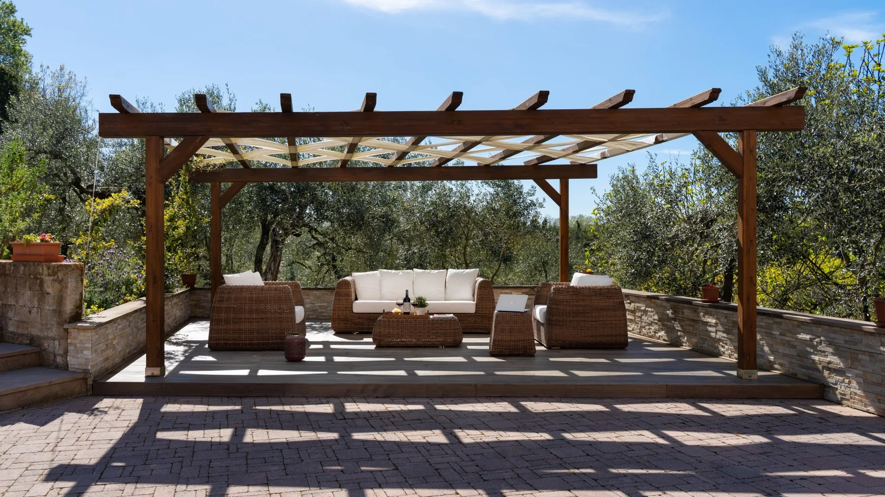Un patio exterior pavimentado con asientos acolchados bajo una pérgola de madera.
