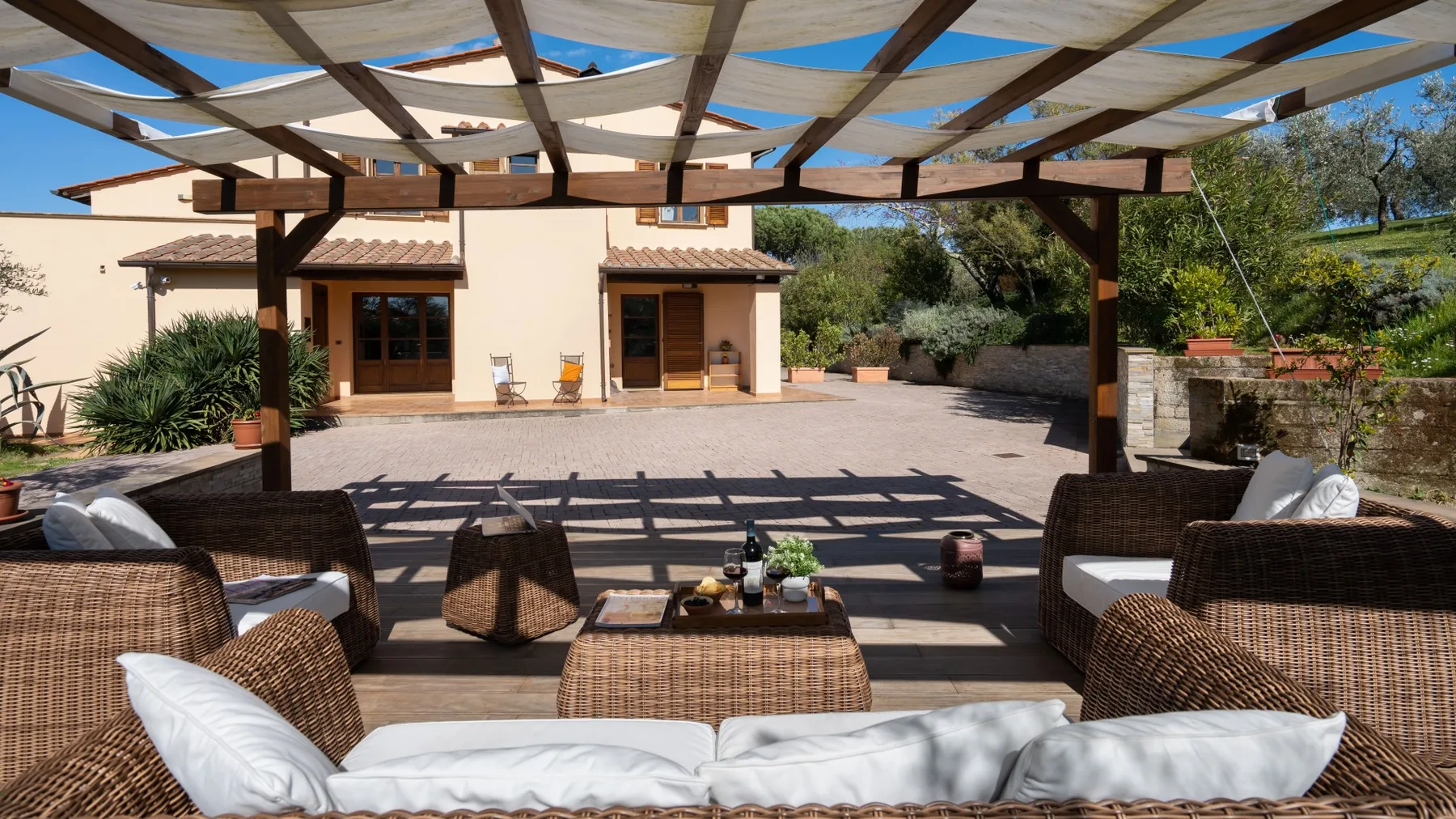 Un patio al aire libre bajo una pérgola de madera, con asientos acolchados frente a la casa.