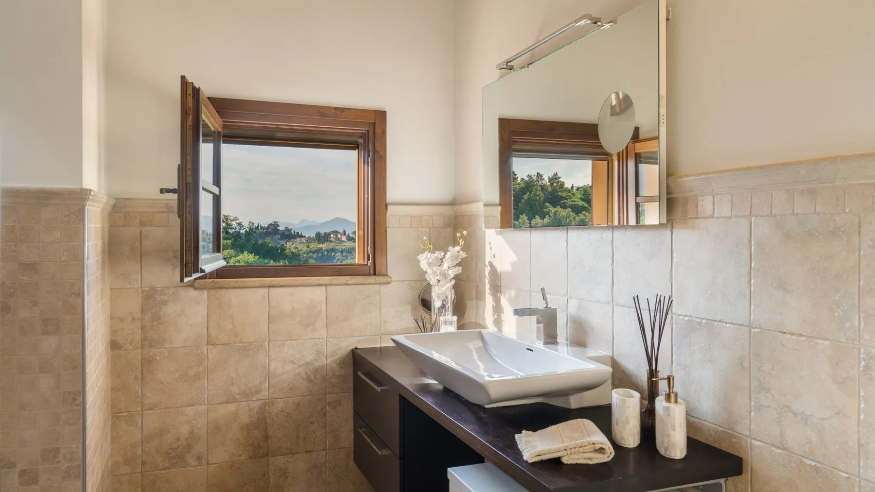 Un tocador de baño con lavabo sobre encimera, espejo y una ventana abierta.