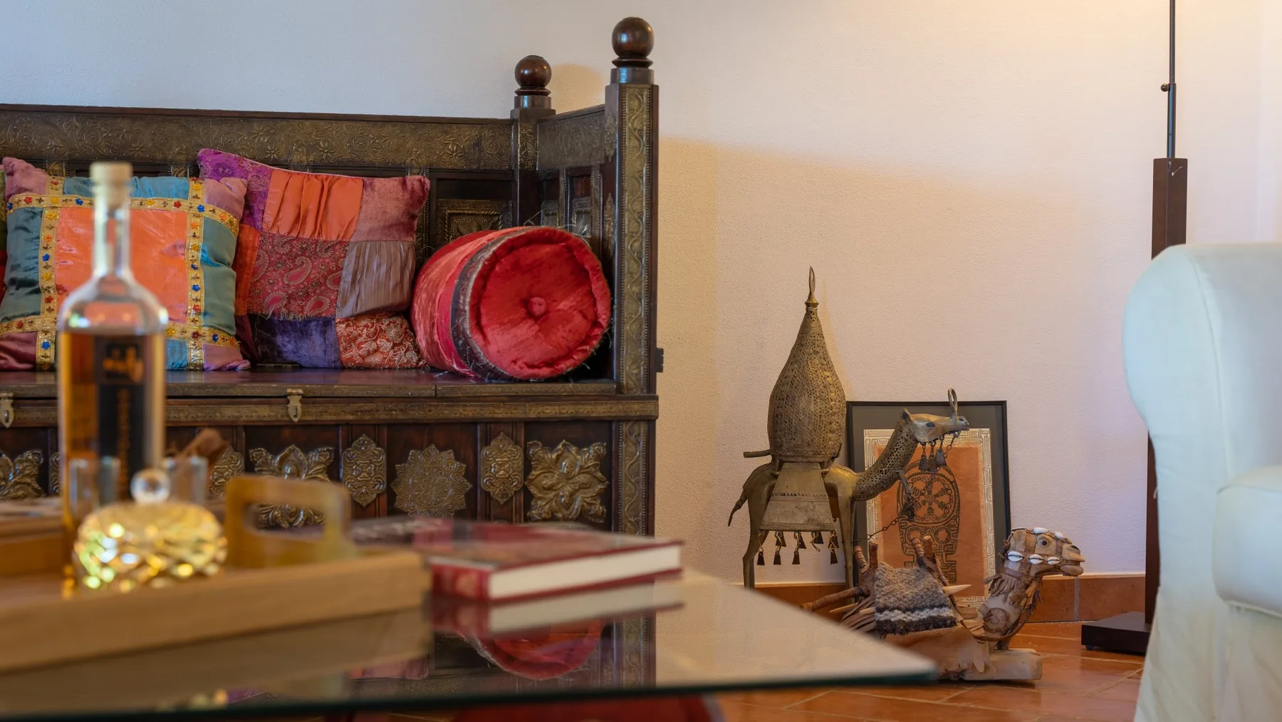 Una sala de estar con un banco de madera oscura, cojines coloridos y una mesa de centro de vidrio.