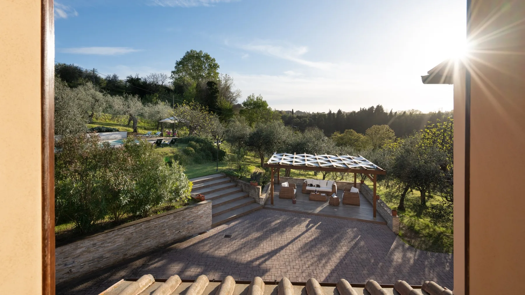 Un patio al aire libre iluminado por el sol da a un jardín con árboles y una pérgola.
