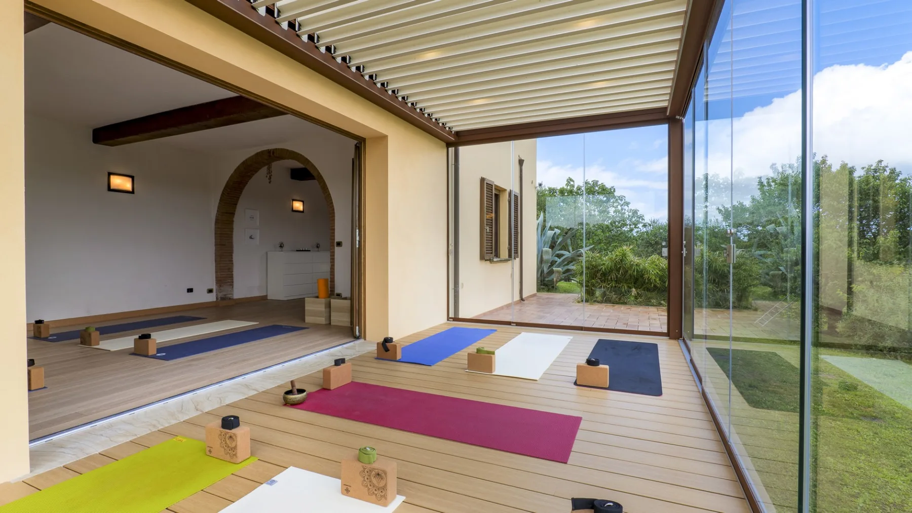 Una amplia sala interior y exterior con colchonetas y paredes de vidrio frente a la vegetación.