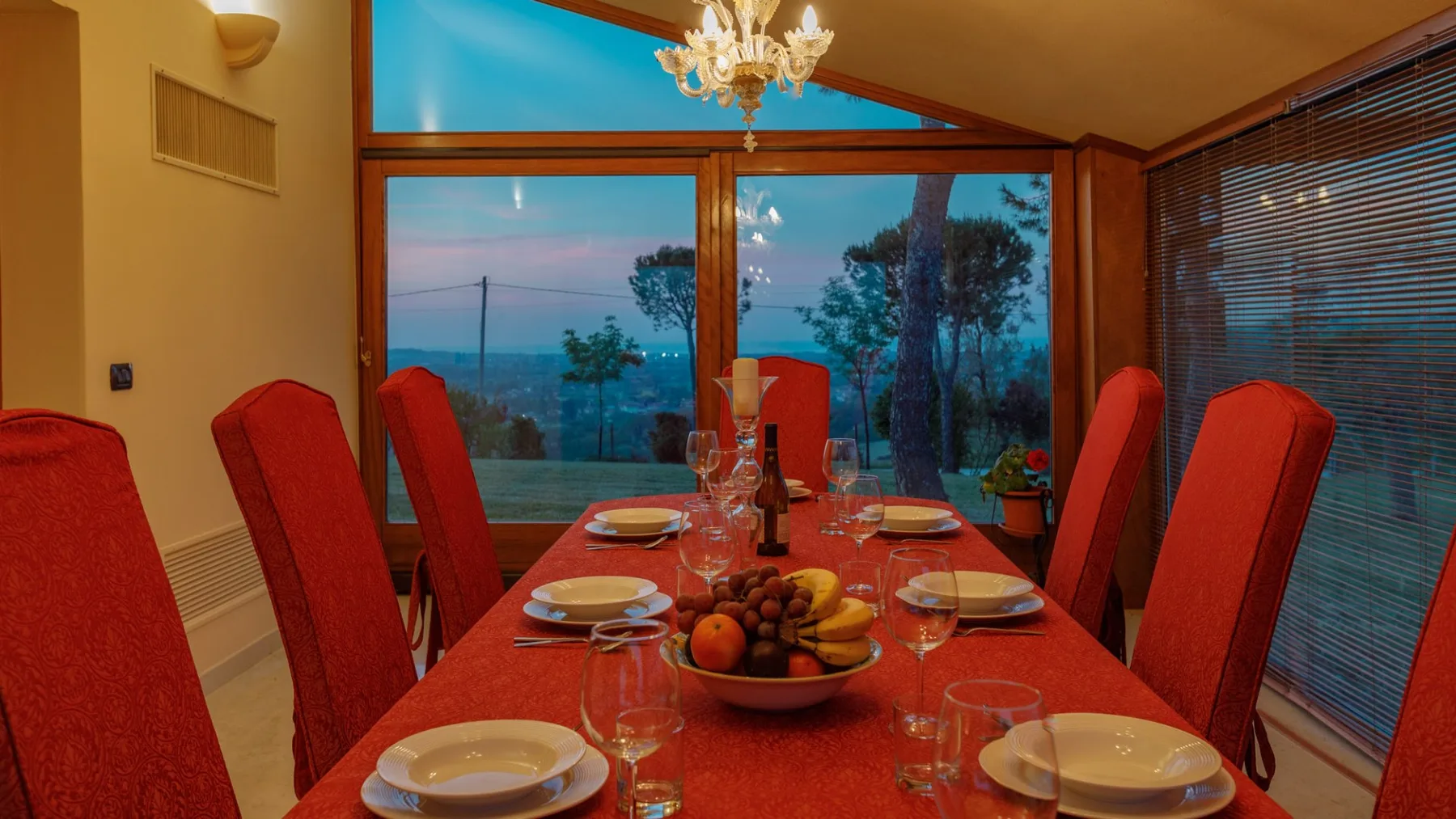 A dining table set with plates and fruit sits beside large windows overlooking trees and water.