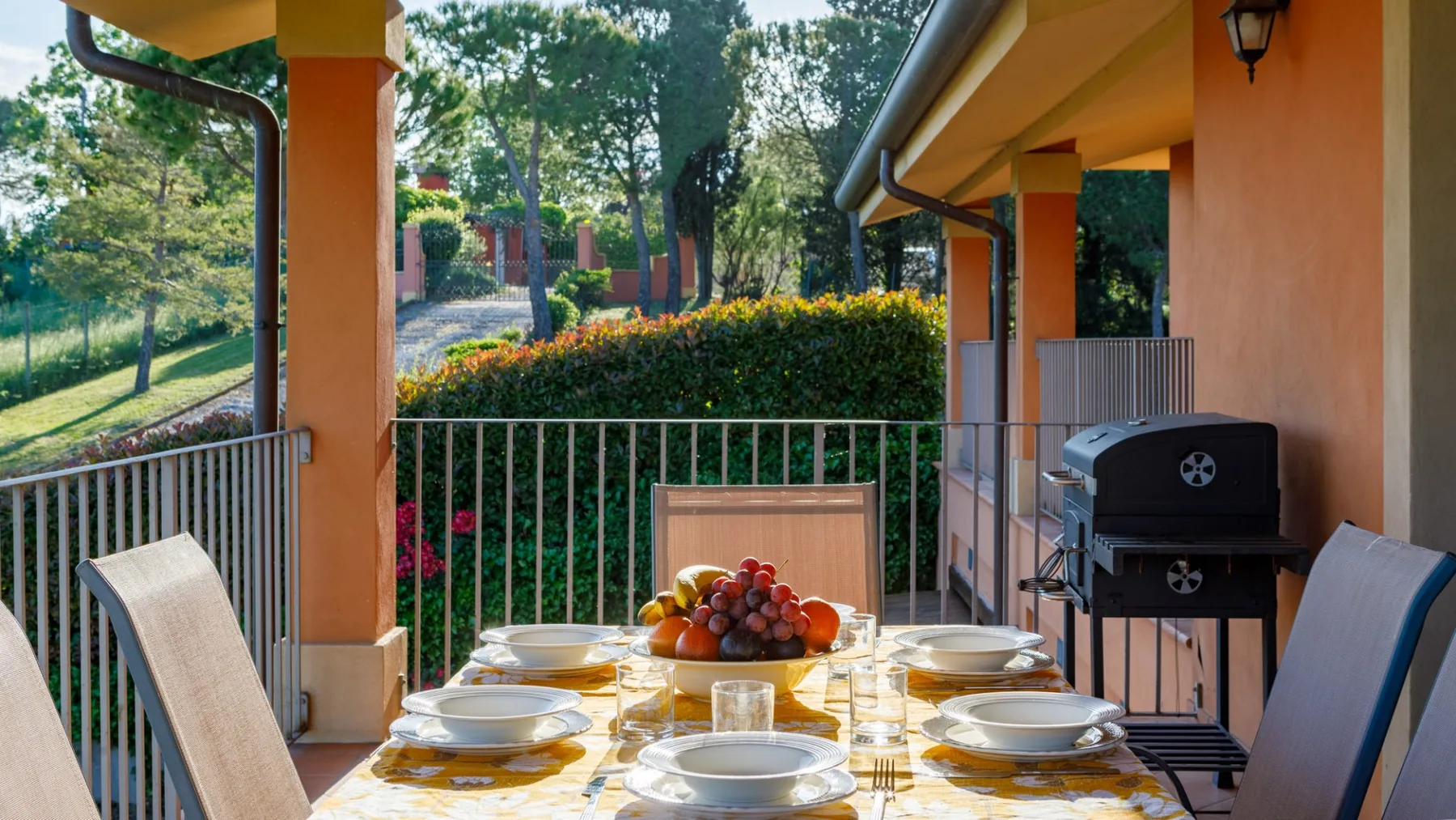 A covered terrace dining area has a set table, metal railing, and barbecue beside flowering hedges.