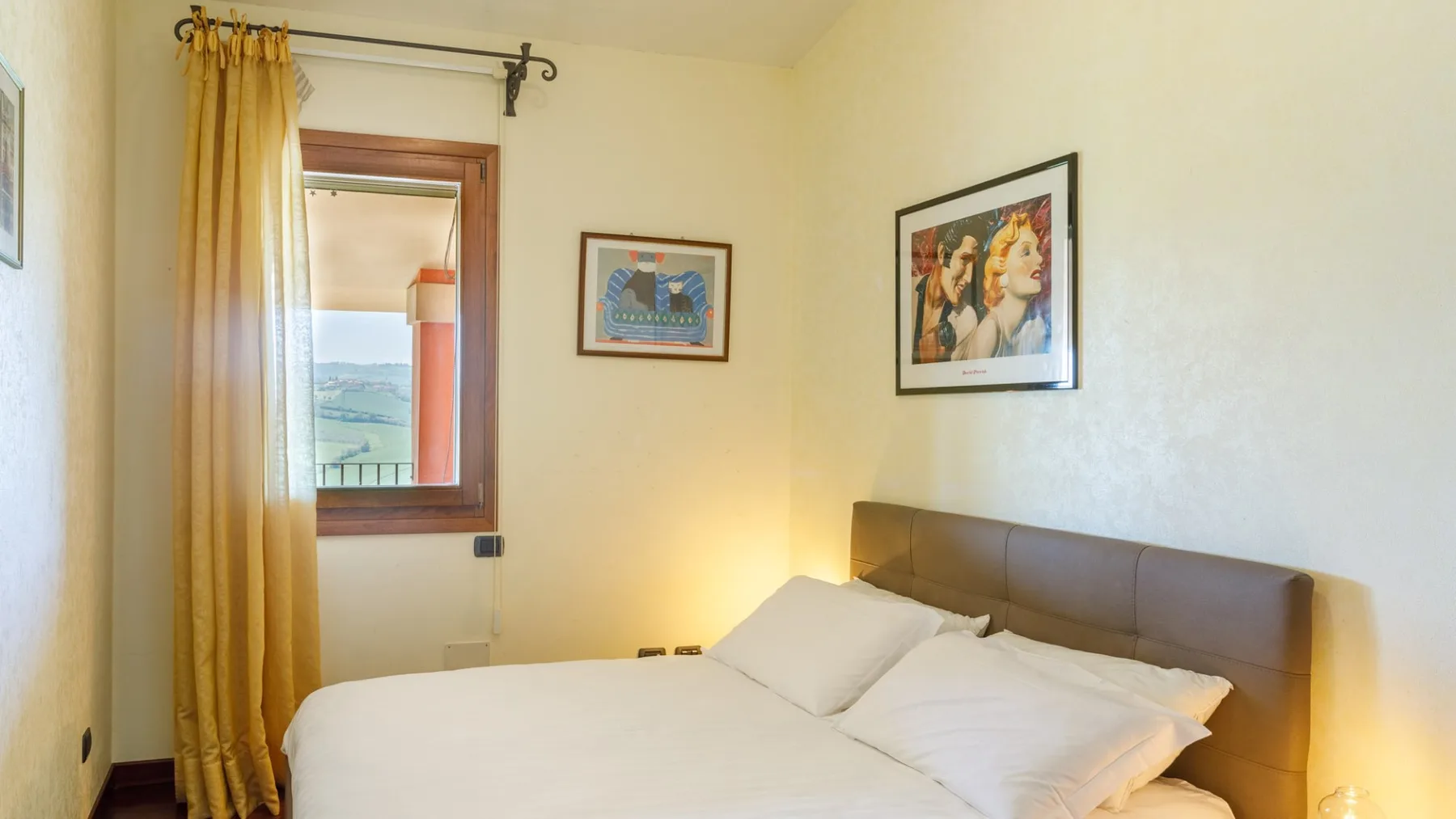 A bedroom with a neatly made bed, brown upholstered headboard, yellow curtains, and framed artwork.