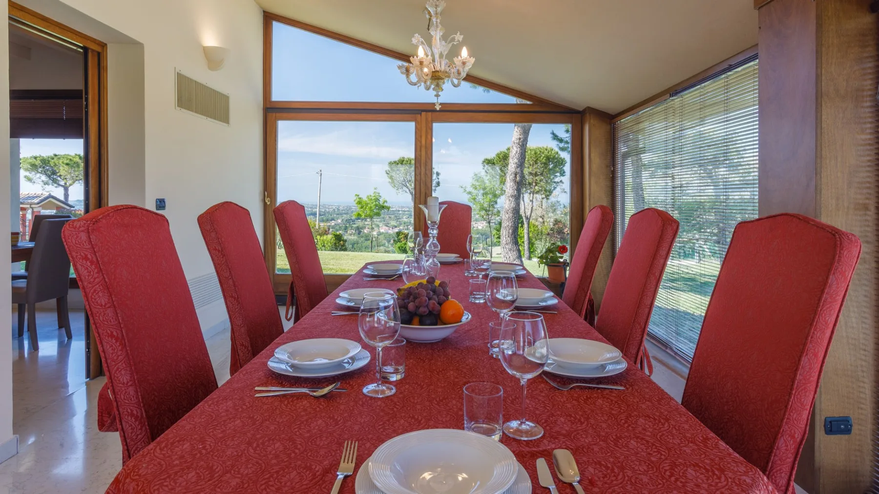 A dining room features a long red table set with plates and glasses, surrounded by red chairs beside large windows.