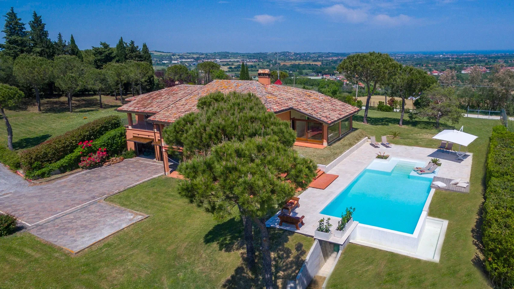 A low tiled-roof house sits beside a bright blue swimming pool, paved patio, and landscaped lawn.