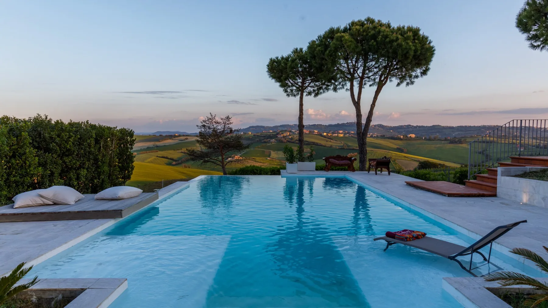 Outdoor swimming pool overlooking rolling green fields, with lounge chairs and tall trees beneath a clear sky.
