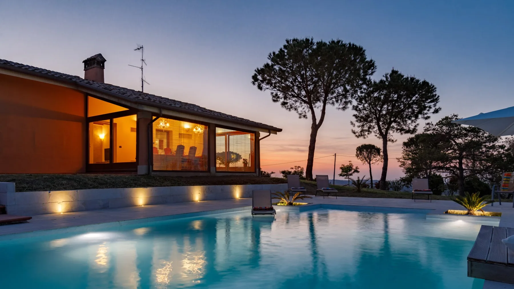An illuminated modern house overlooks a swimming pool, with trees and a sunset sky in the background.