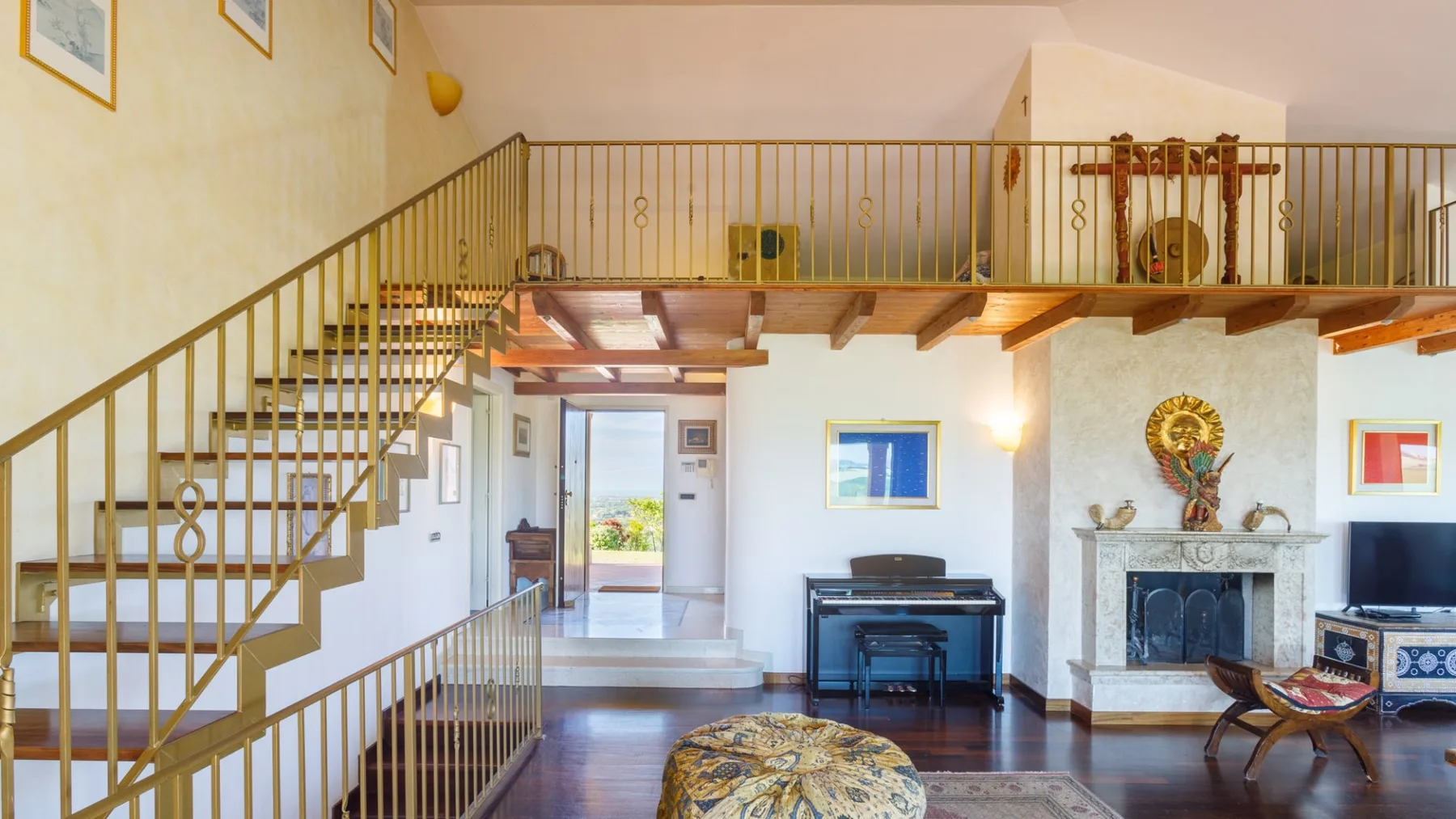 A spacious living room features a brass-railed staircase, an upright piano, and a decorative fireplace.