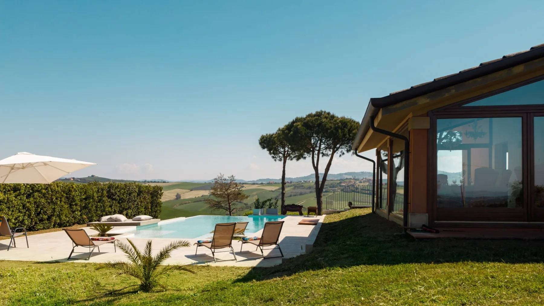 A sunny outdoor pool beside a modern house, with patio chairs, an umbrella, and rolling hills beyond.