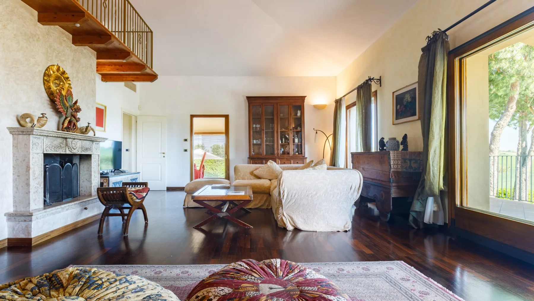 A bright living room with a stone fireplace, wooden staircase, sofa, and large windows overlooking greenery.