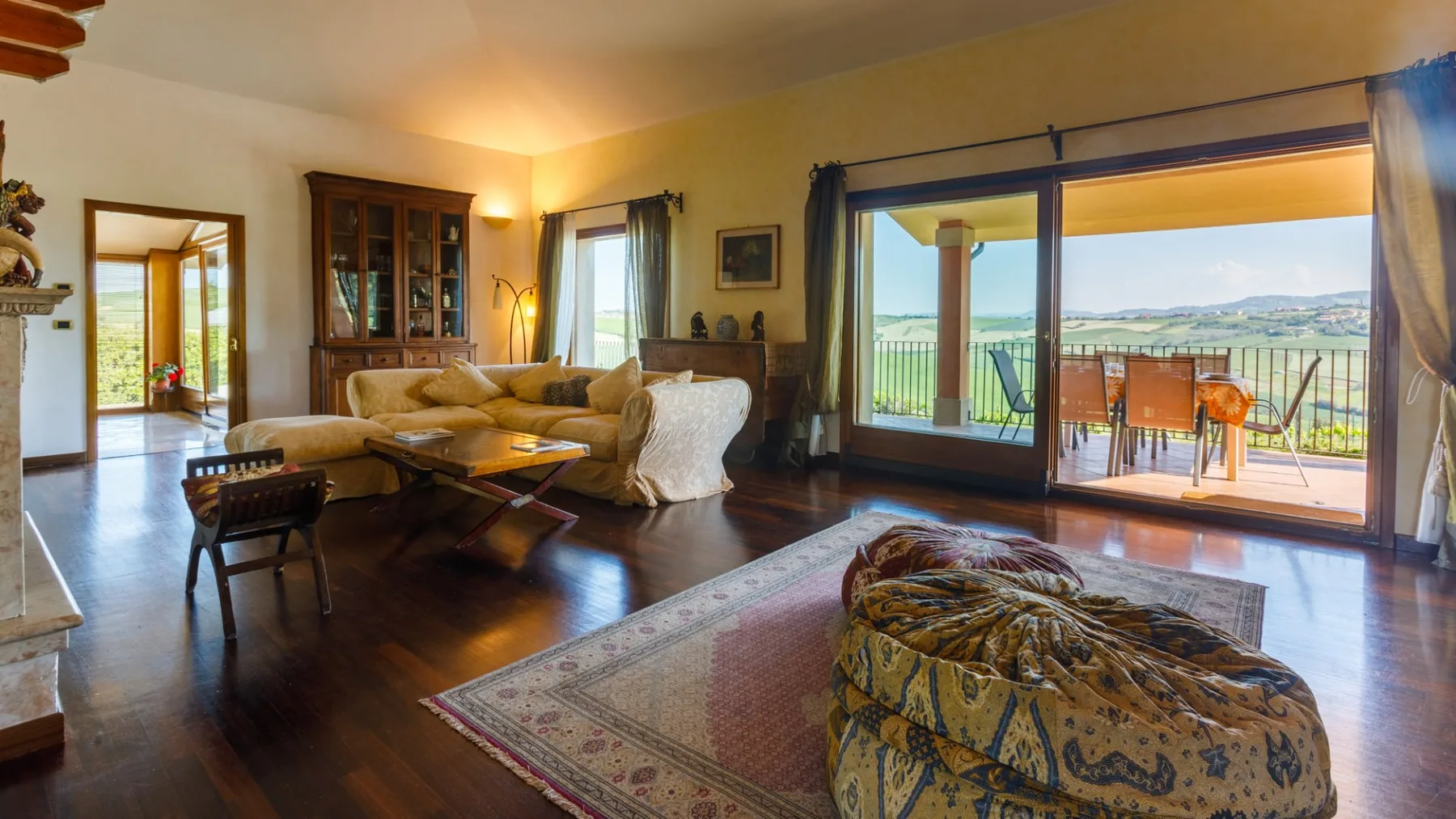 A bright living room has a sectional sofa, glass-front cabinets, and sliding doors opening onto a balcony.
