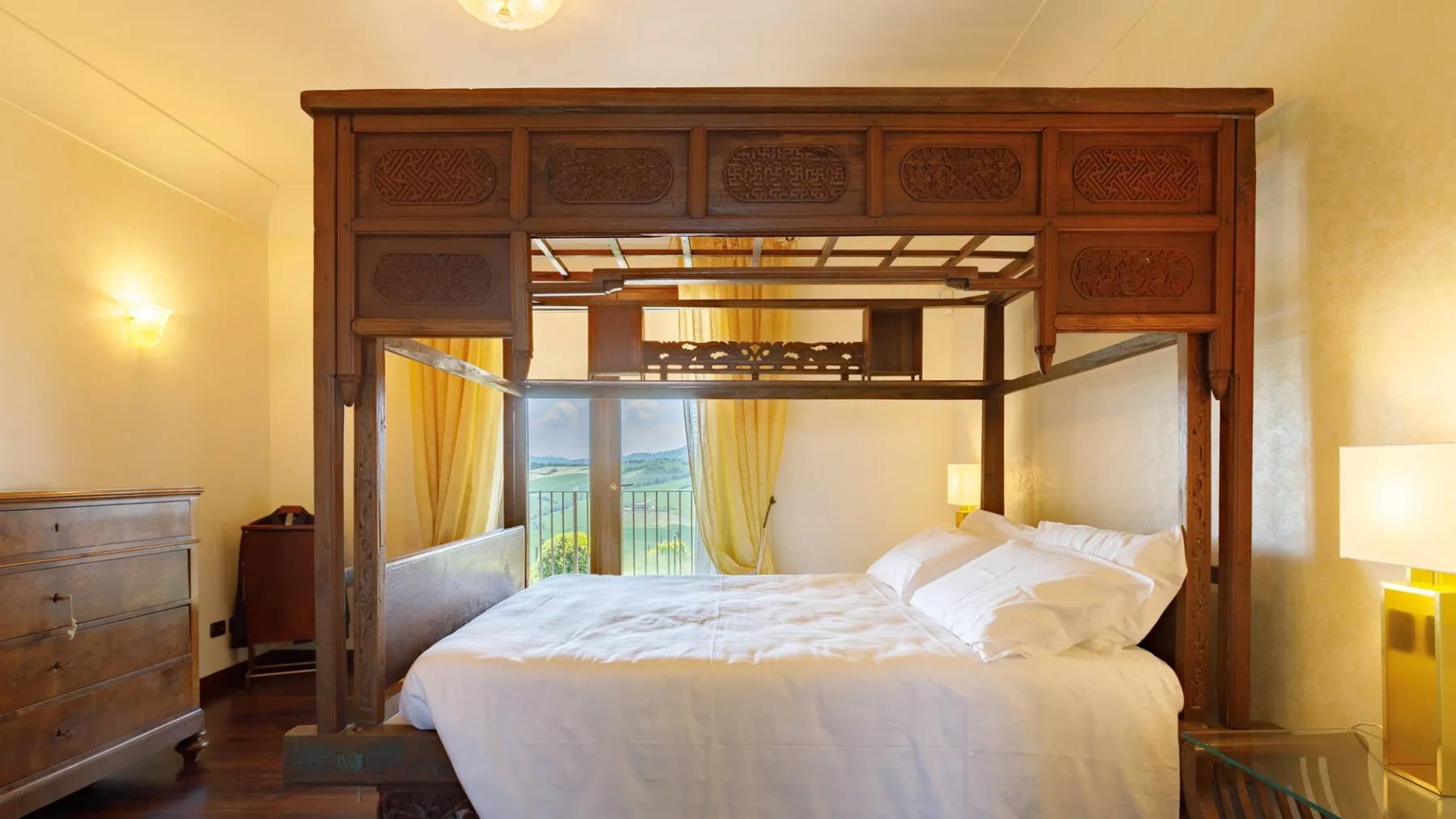 A bedroom with a large carved wooden four-poster bed, white bedding, and yellow curtains by the windows.