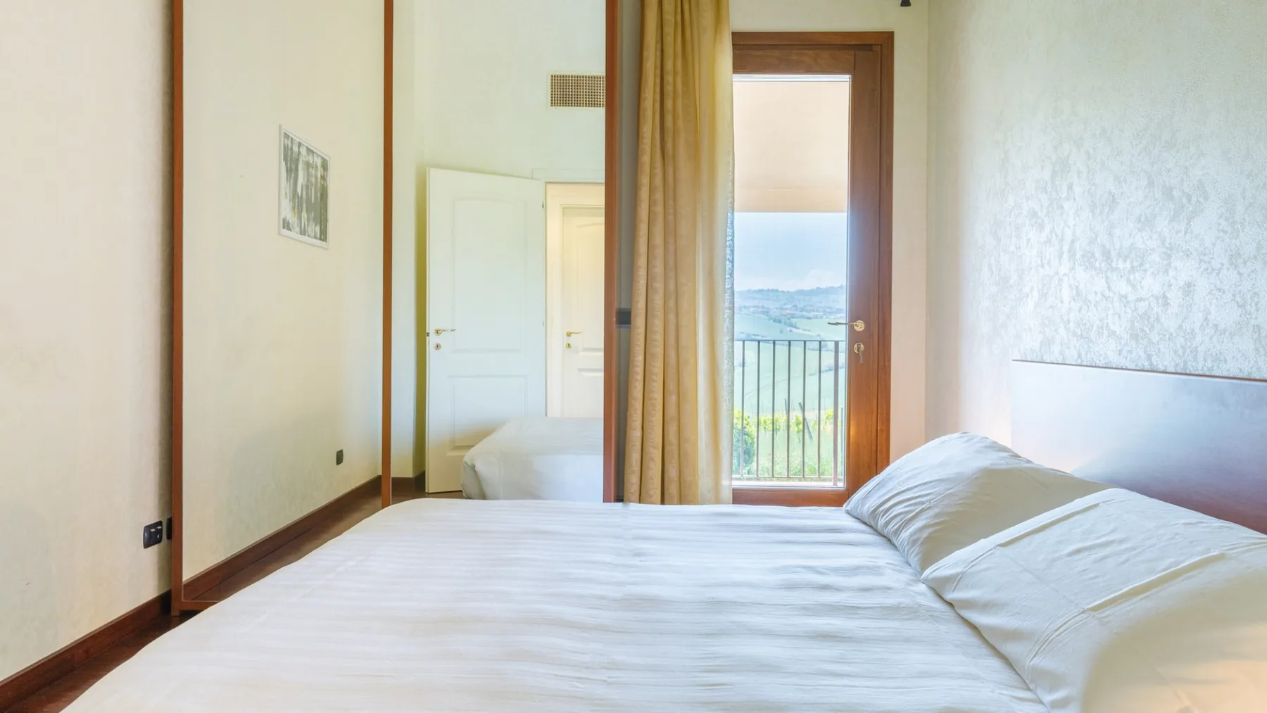 A bedroom with a neatly made bed, a curtained balcony door, and a view of the water.
