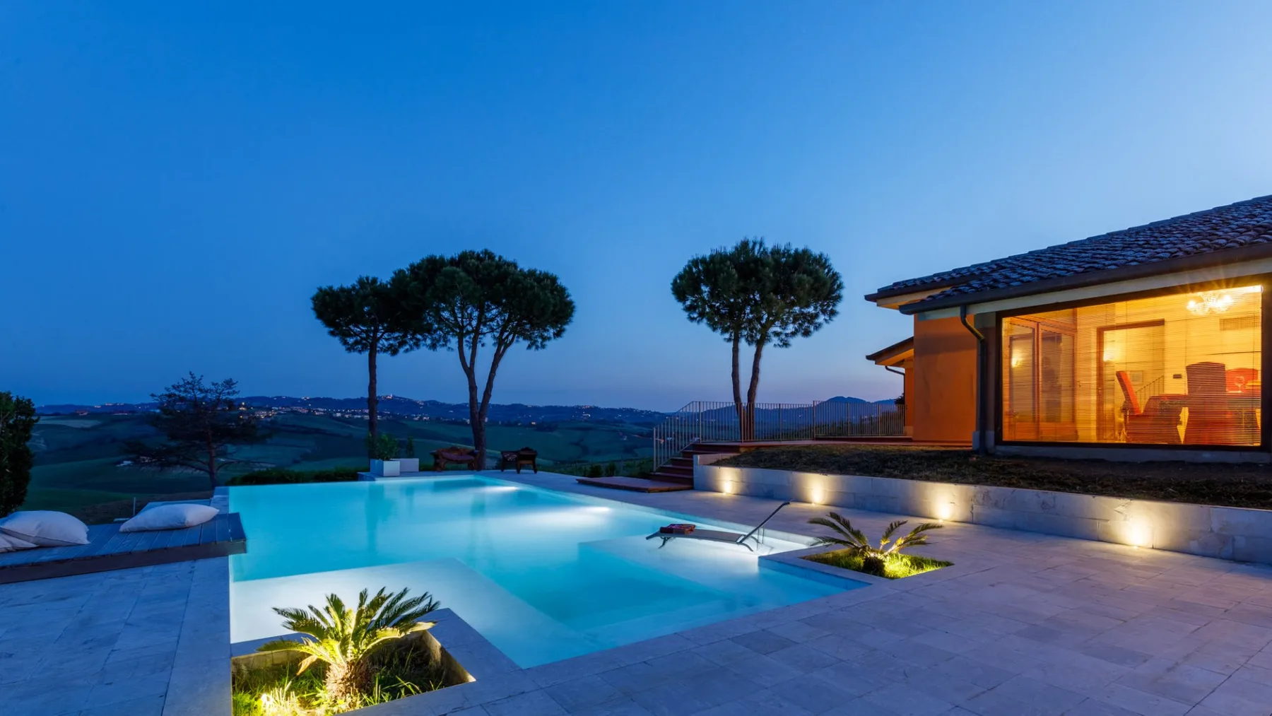 An illuminated swimming pool and tiled terrace sit beside a glass-fronted house at dusk.