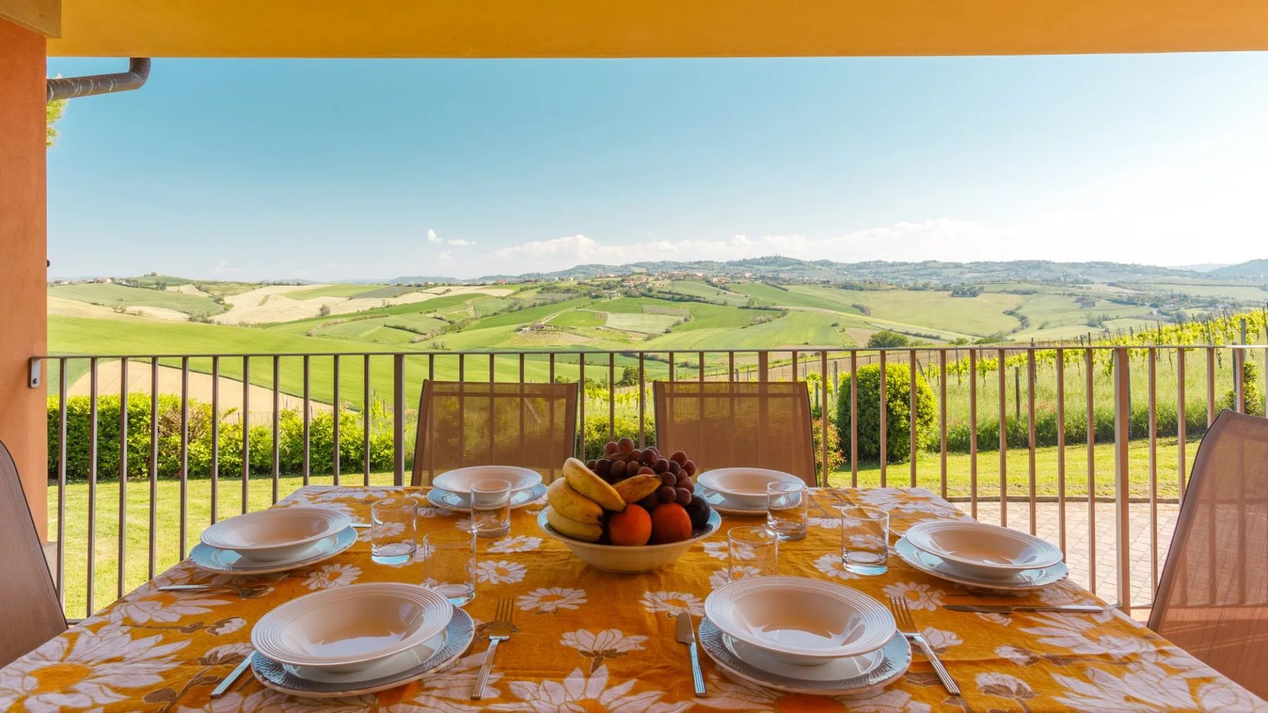 A covered terrace dining table is set with plates and fruit, overlooking green rolling hills.