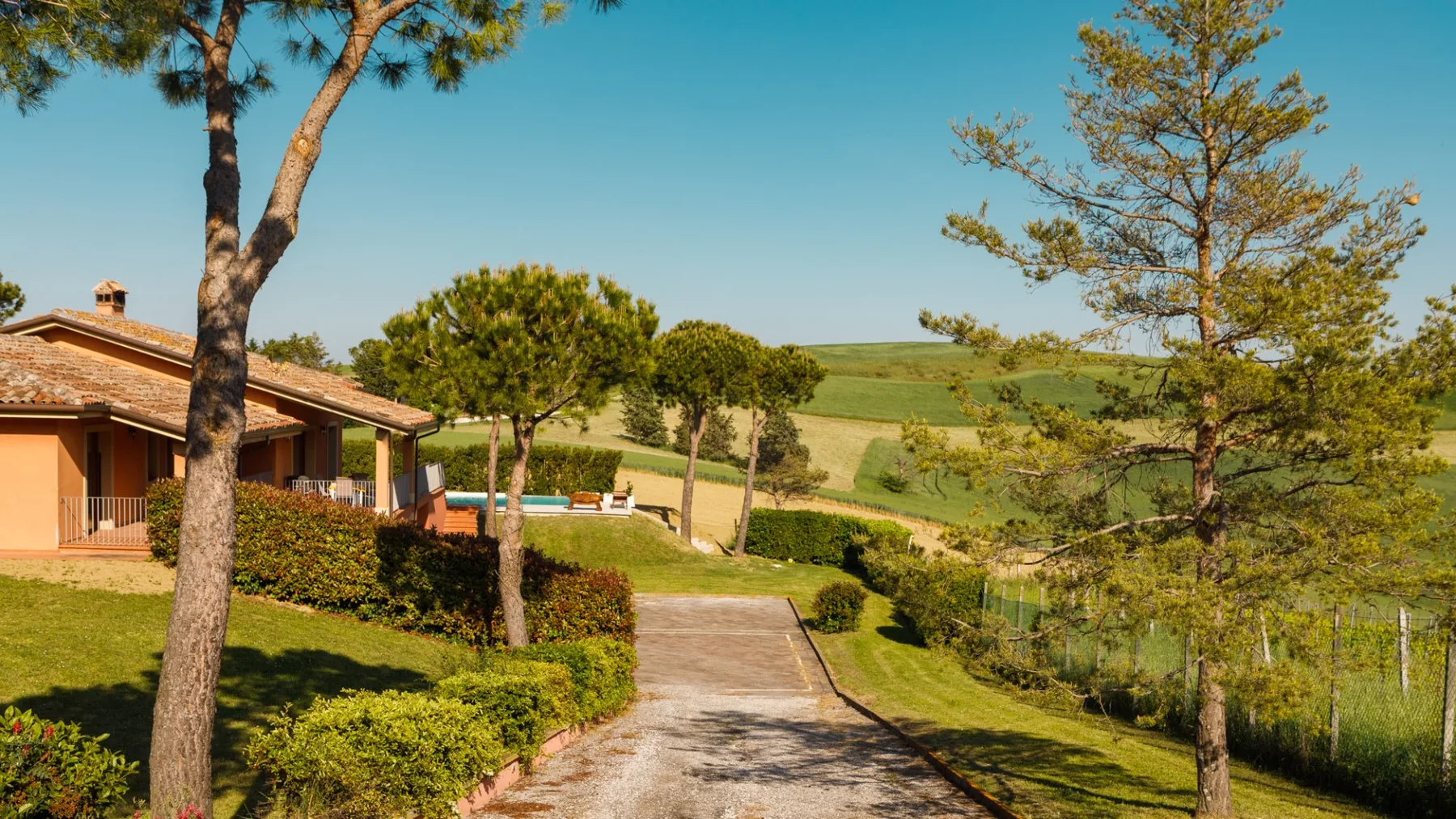 A country house stands beside a paved driveway, with trimmed hedges, trees, and rolling green fields.