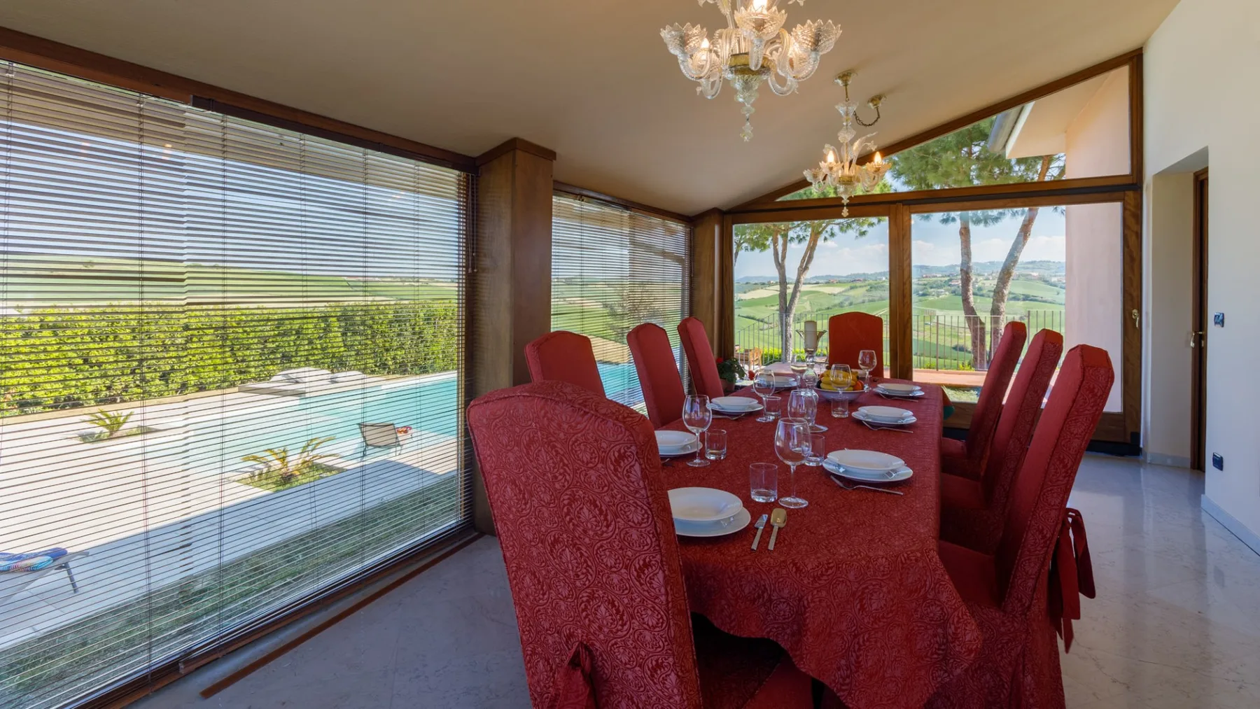 A formal dining room has a long table set with red linens, surrounded by upholstered chairs and large windows.