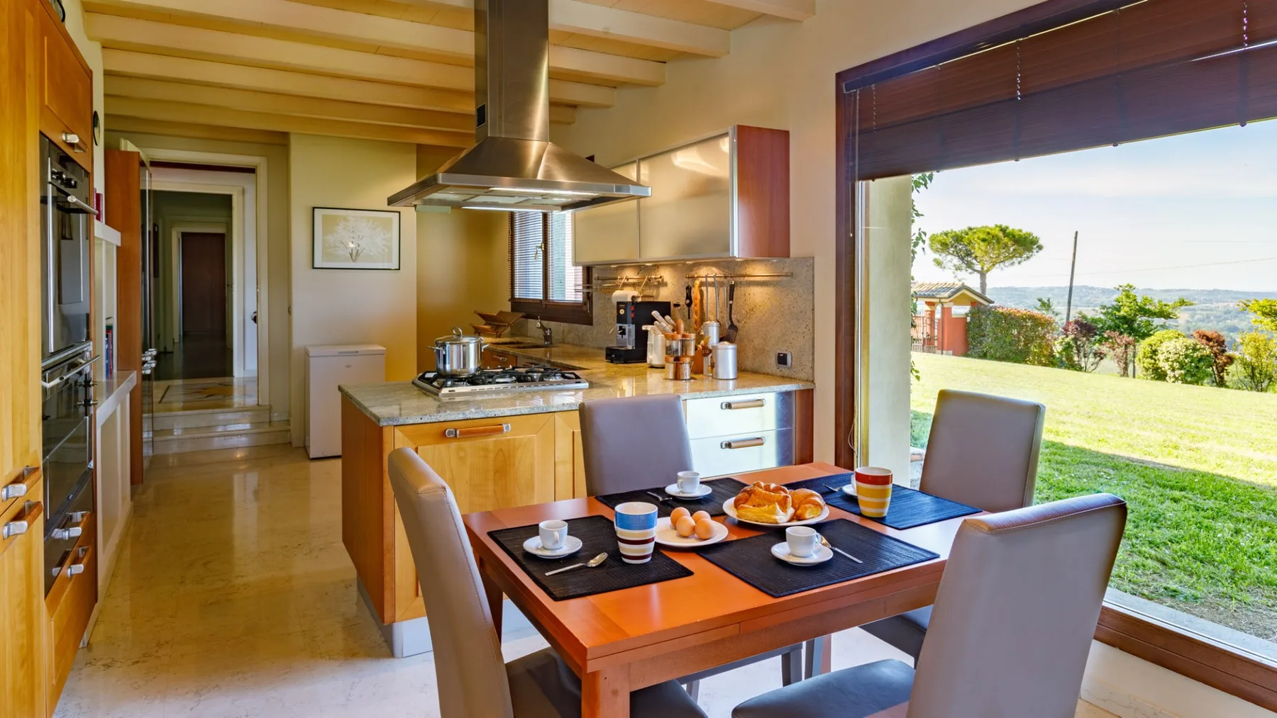 A dining table with place settings sits beside a bright kitchen, with a large window overlooking trees and water.