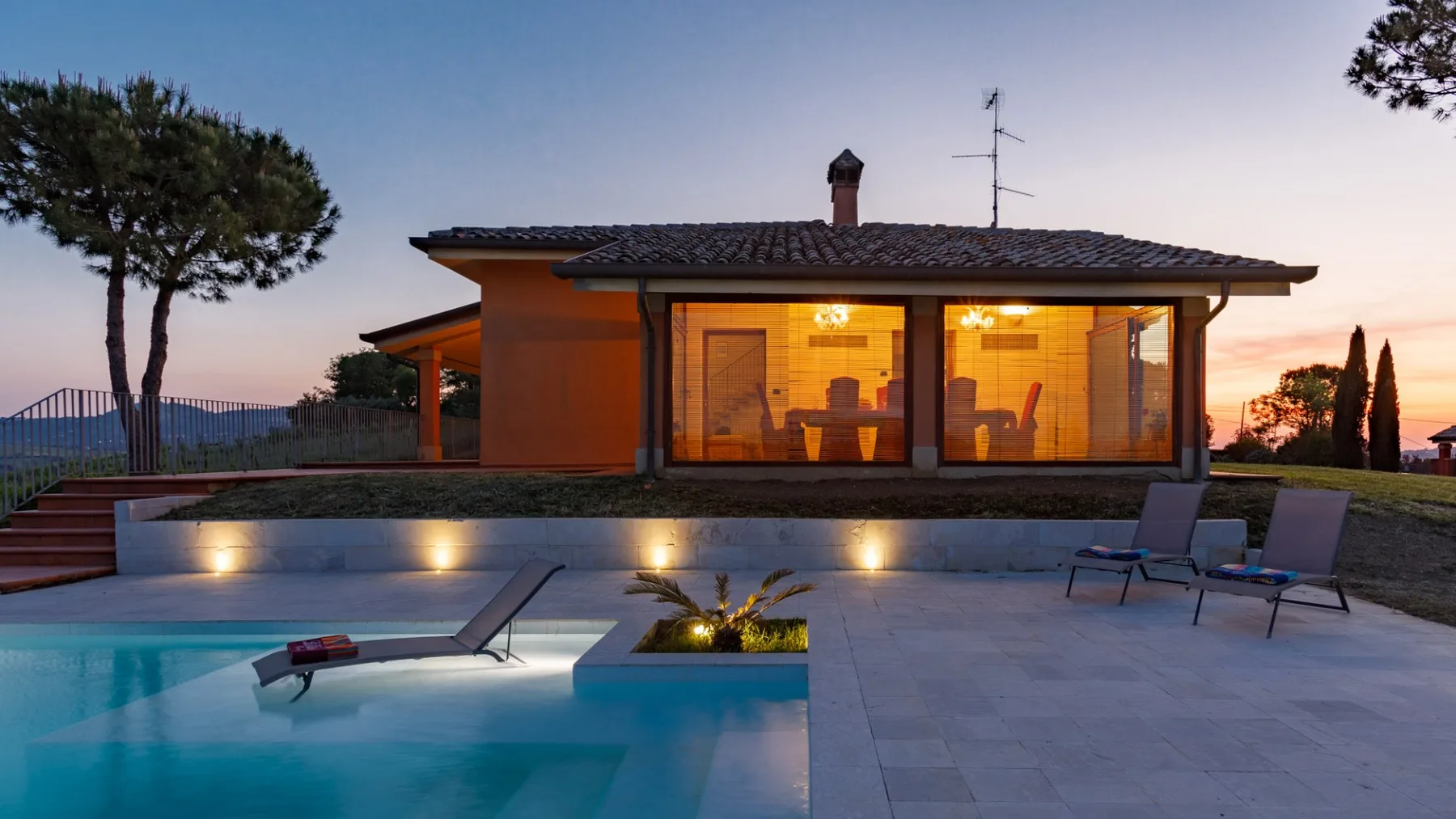 A modern house with warmly lit glass doors overlooks a swimming pool and paved patio at dusk.