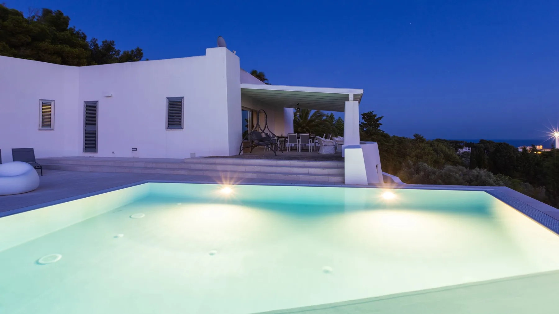 Una piscina exterior iluminada está junto a un edificio blanco moderno al anochecer.