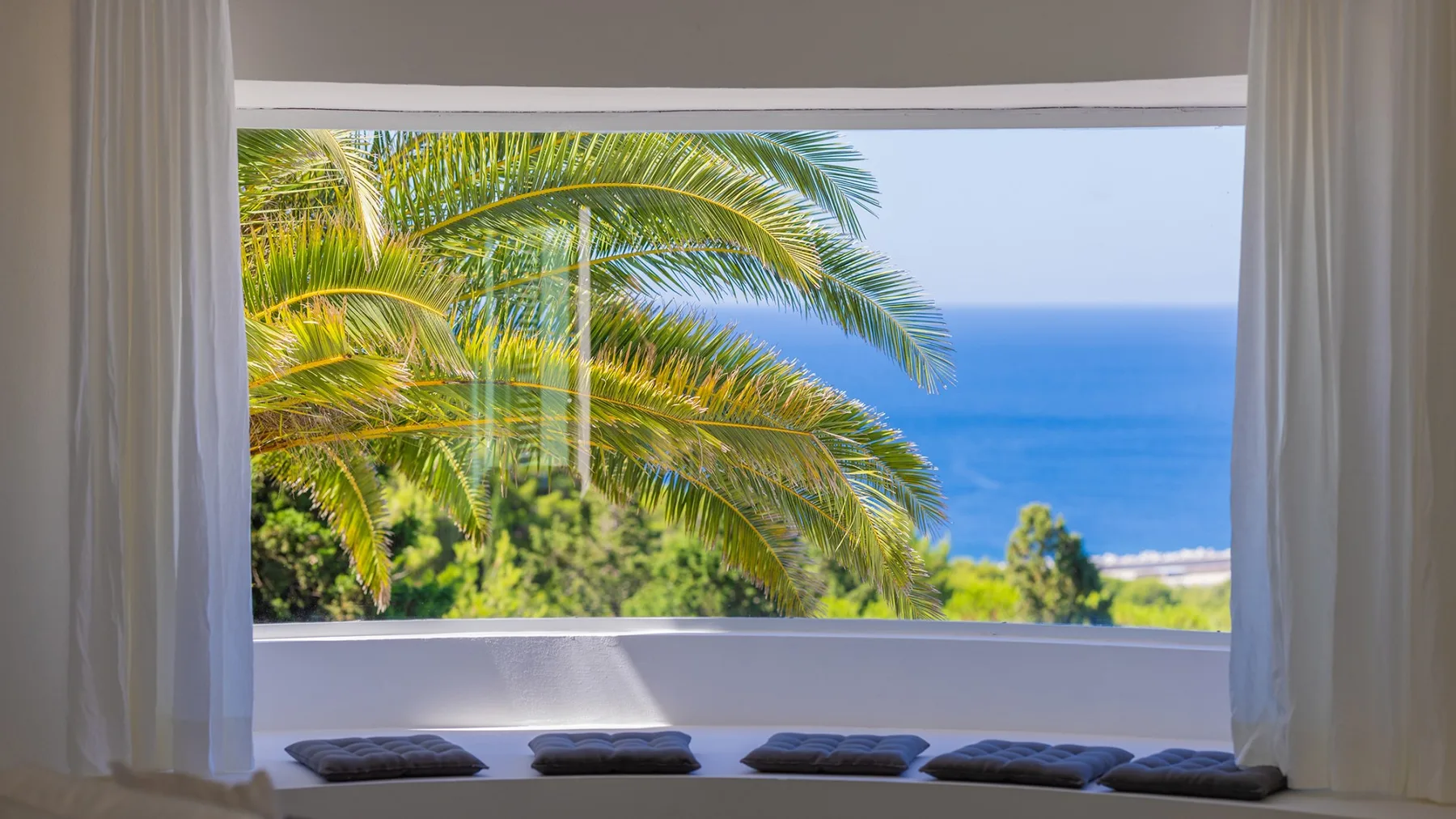 Una ventana amplia enmarca palmeras y una vista del mar azul.