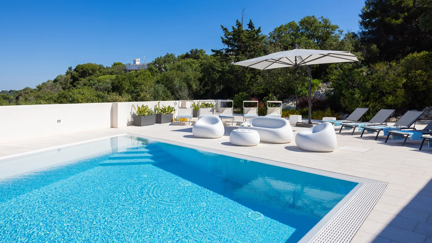 Una piscina al aire libre junto a una terraza soleada con tumbonas y una sombrilla grande.