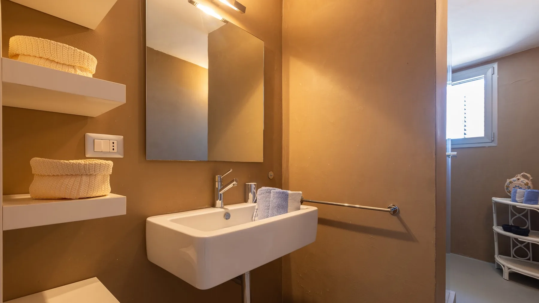 Un baño con lavabo de pared, espejo y estantes.