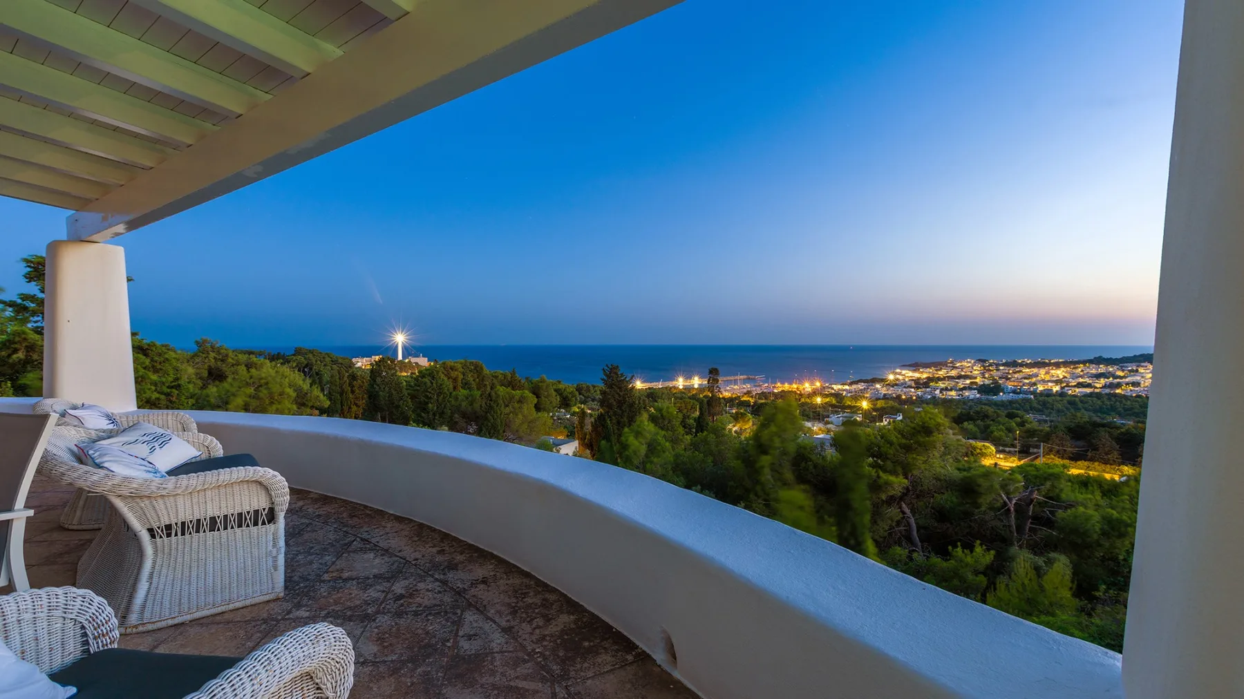 Una terraza cubierta da a árboles, luces lejanas de la ciudad y el mar al anochecer.