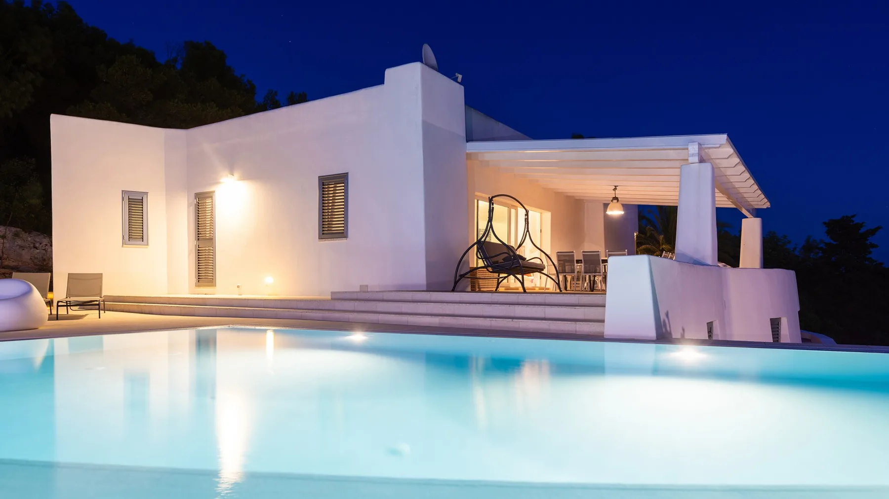 Una casa blanca moderna está junto a una piscina iluminada al anochecer.