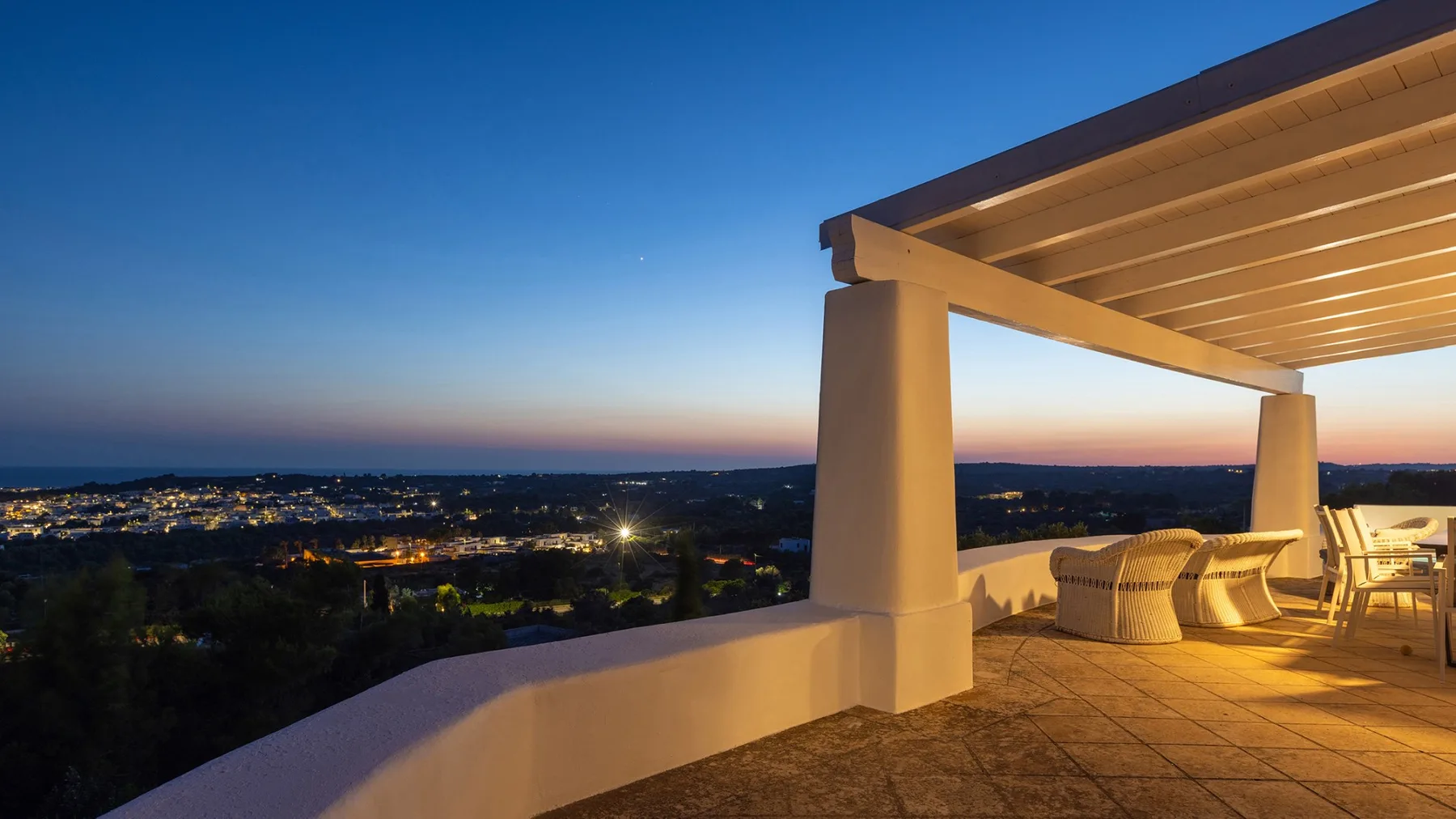 Una terraza cubierta domina una ciudad al atardecer, con asientos bajo una pérgola.