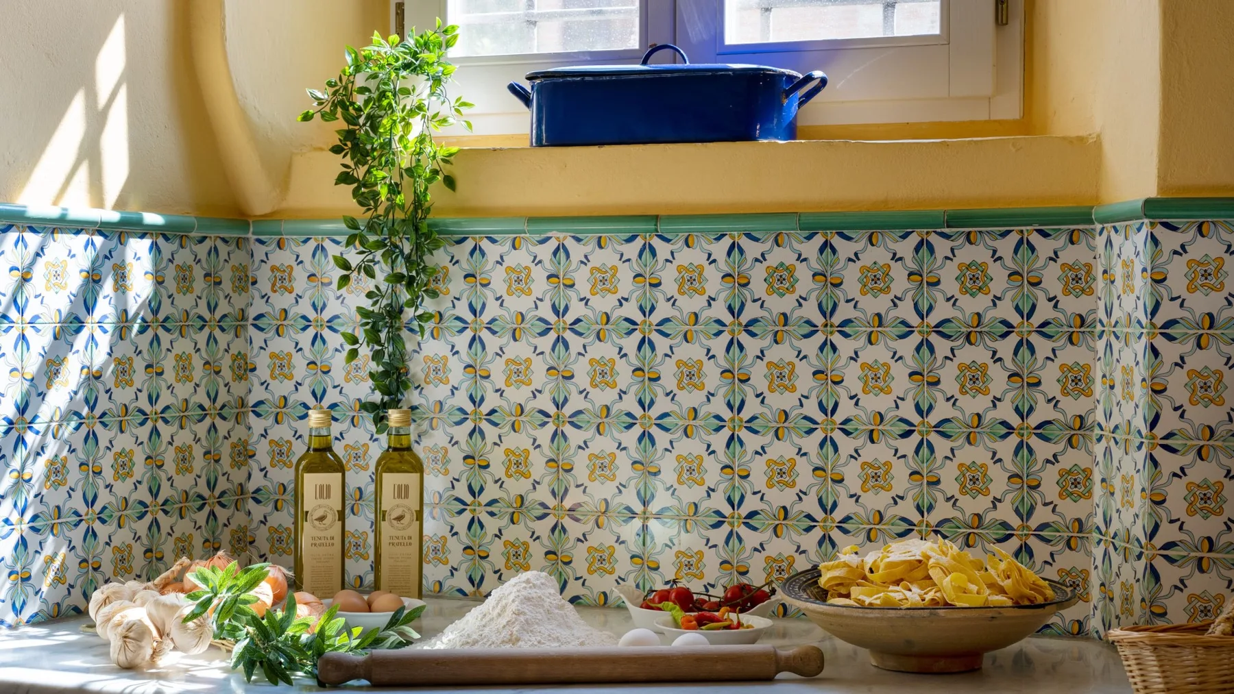 A tiled kitchen counter displays vegetables, herbs, and bowls beneath a window.