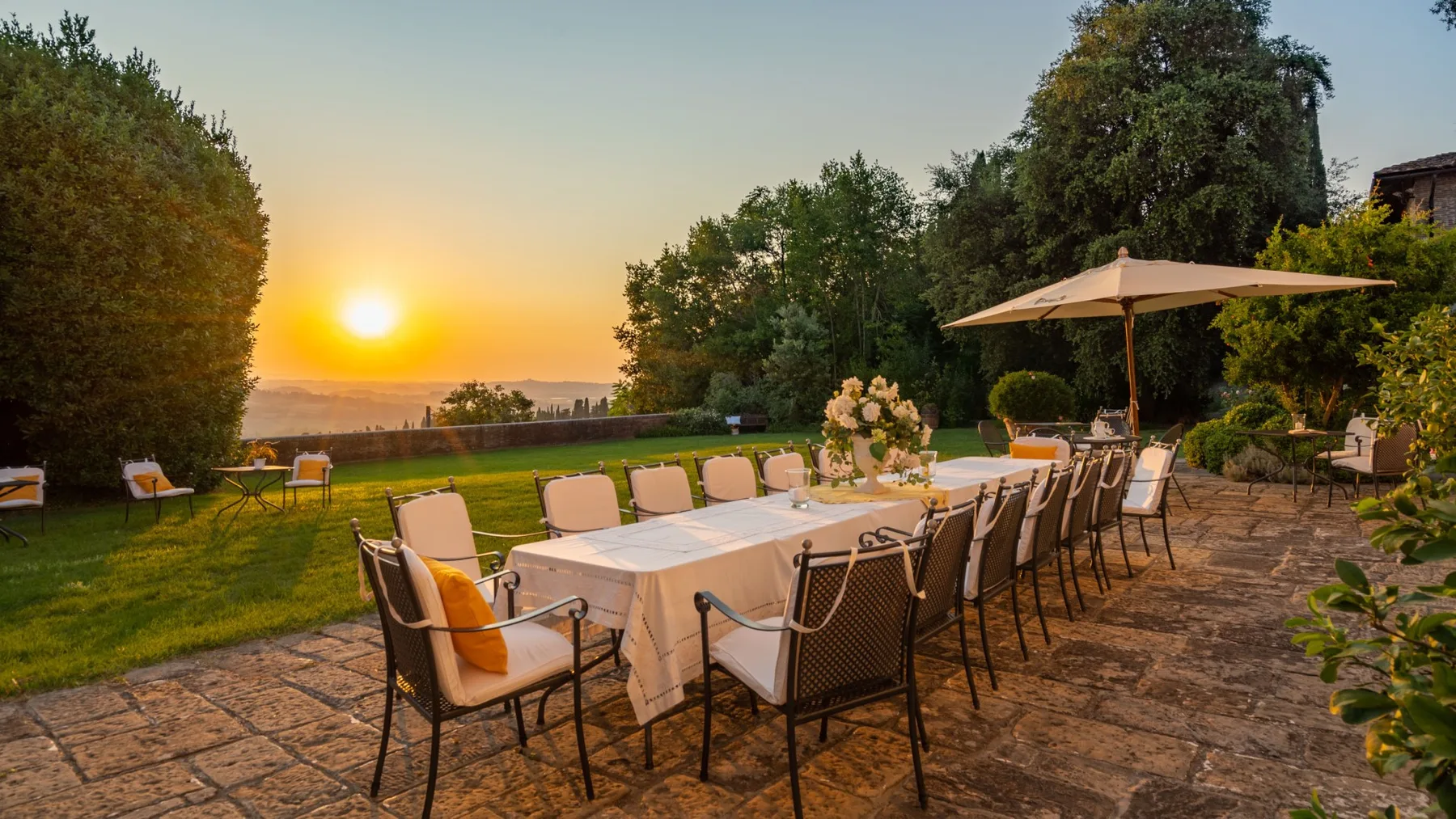 An outdoor dining terrace with a long table, chairs, and a large umbrella at sunset.