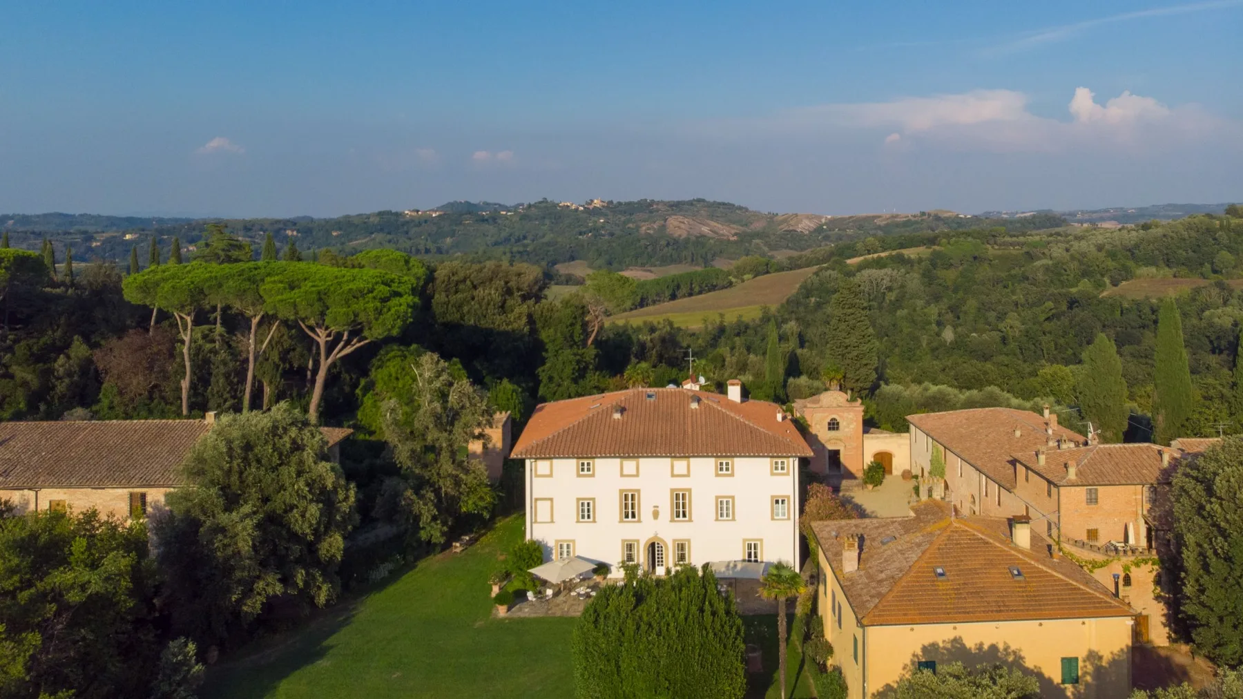 A countryside estate with pale buildings, terracotta roofs, gardens, and rolling hills in the distance.
