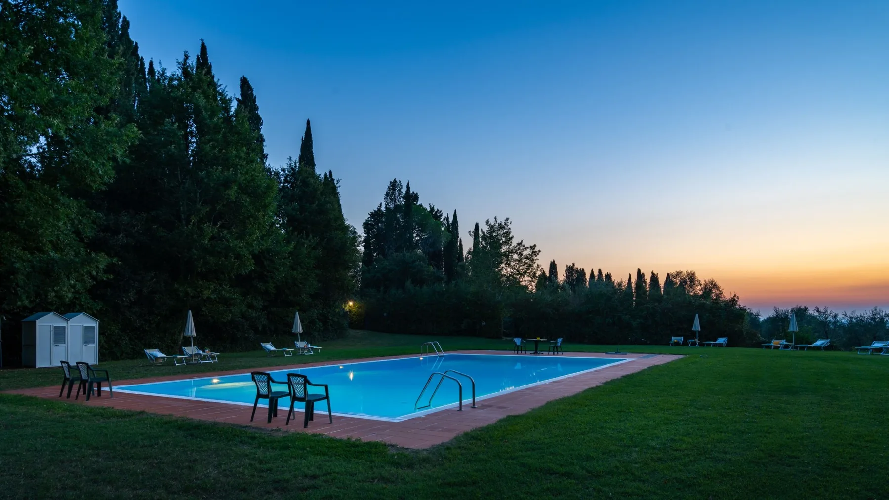 An outdoor swimming pool sits beside a grassy lawn at dusk.