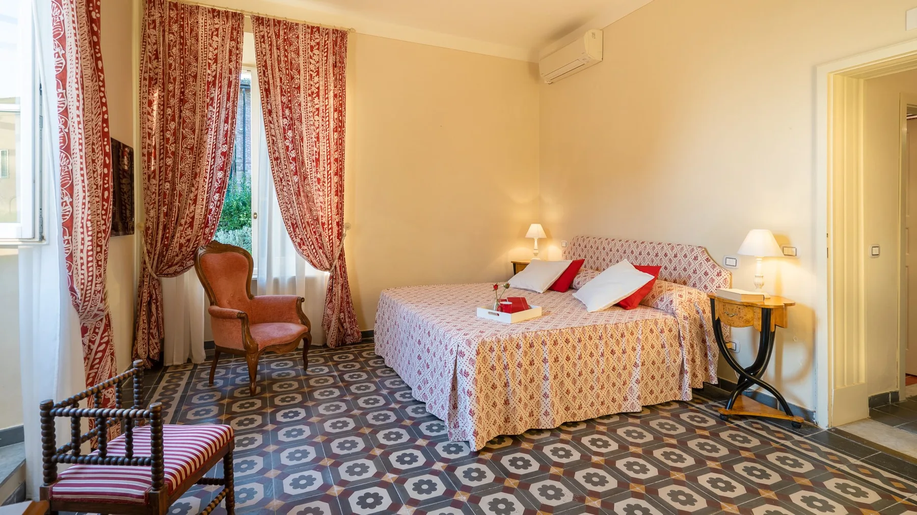 A bedroom with a patterned bed, a small table, and tall curtained windows.